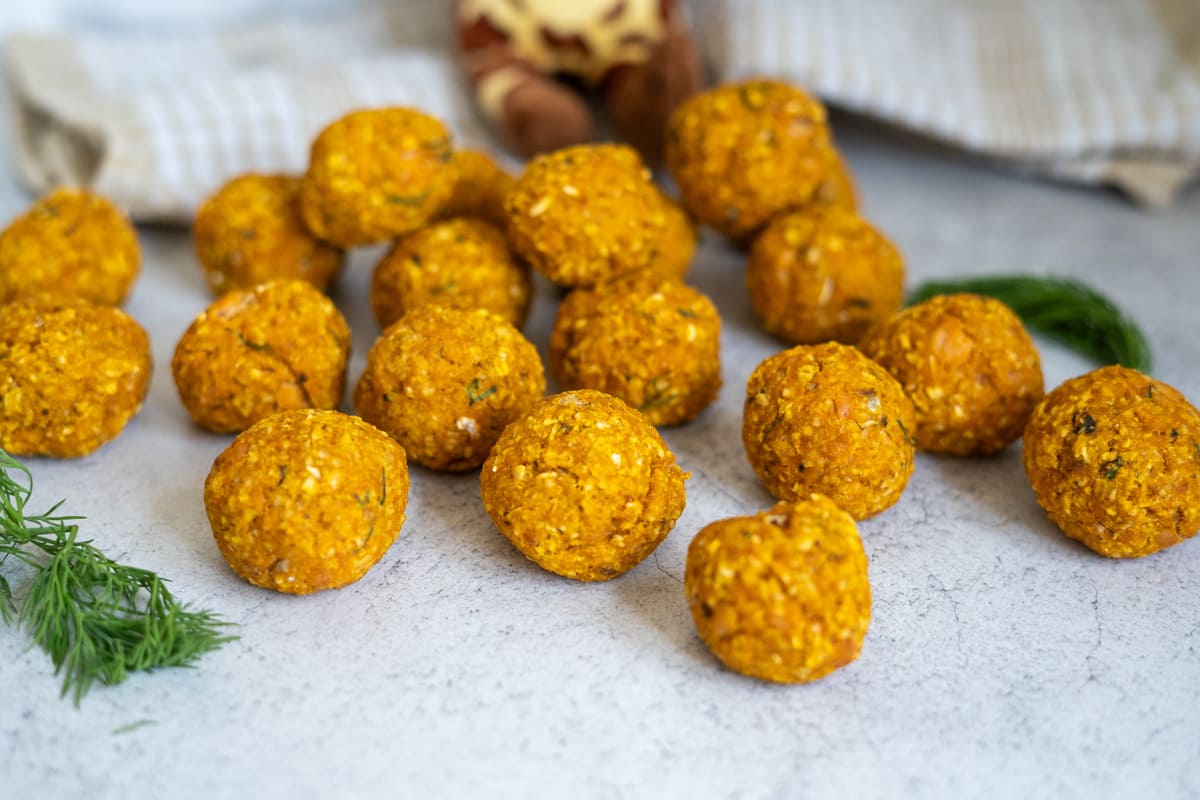 Several orange-coloured falafel balls are arranged on a light-coloured surface with some dill sprigs nearby and a chequered cloth in the background.