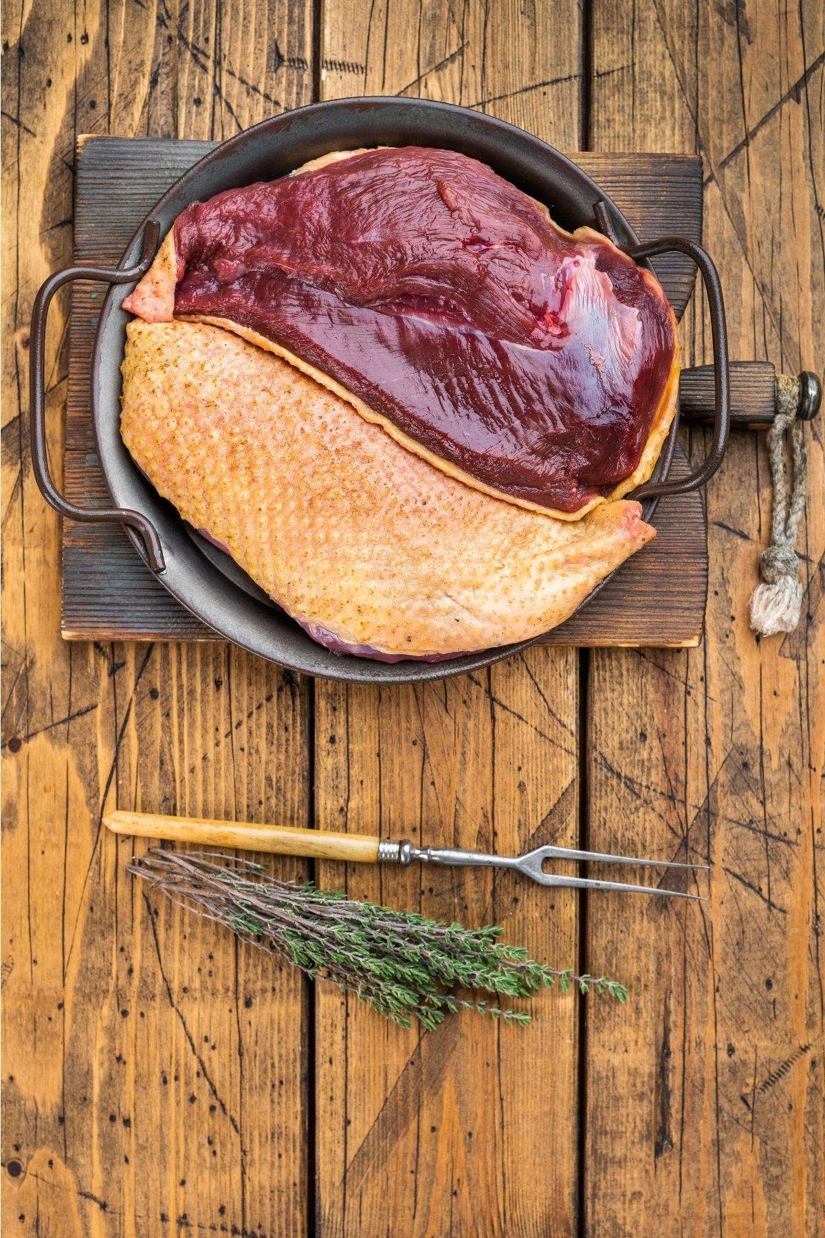 Raw duck breast with skin on is placed in a pan on a wooden board, with a carving fork and herbs beside it; if you're wondering "can dogs eat duck," this scene showcases its preparation on a rustic table.