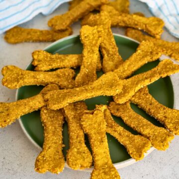 A plate filled with homemade bone-shaped dog biscuits, golden brown in colour, on a light surface.