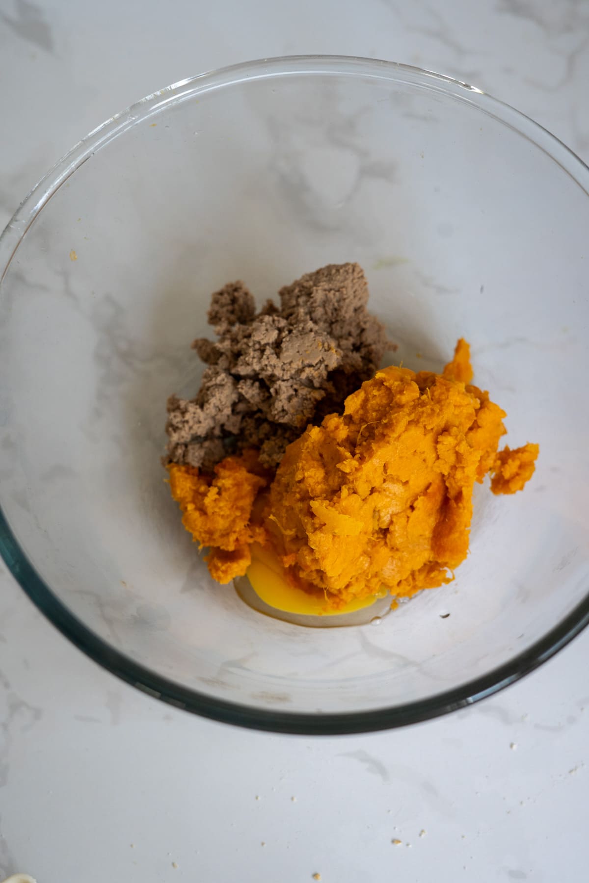 A glass bowl containing minced meat, mashed sweet potato, and a raw egg on a white surface.