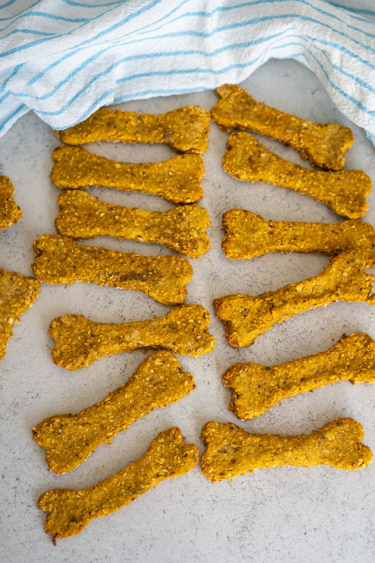 Homemade dog biscuits shaped like bones are arranged on a light surface next to a blue and white striped cloth, resembling classic duck liver dog treats.