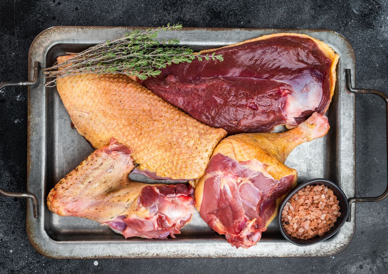 A metal tray with raw duck meat cuts, a bunch of herbs, and a small bowl of pink Himalayan salt on a dark background.