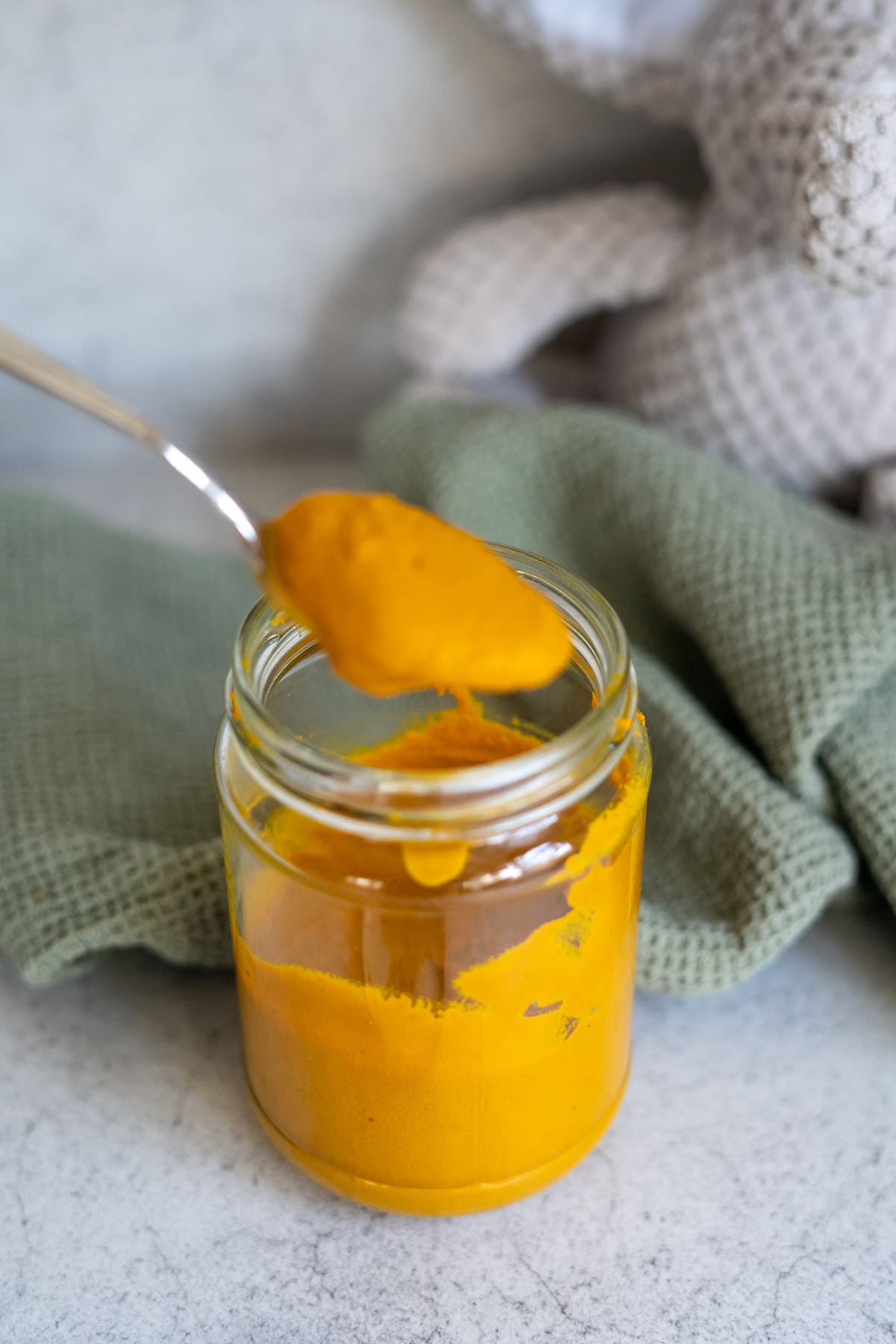 A spoon holding a dollop of thick yellow paste above a glass jar filled with the same paste, with a textured cloth and soft toy in the background.