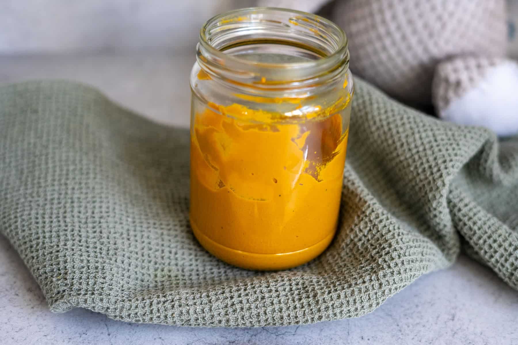 A glass jar filled with bright yellow turmeric paste sits on a textured green cloth, with a blurred background.