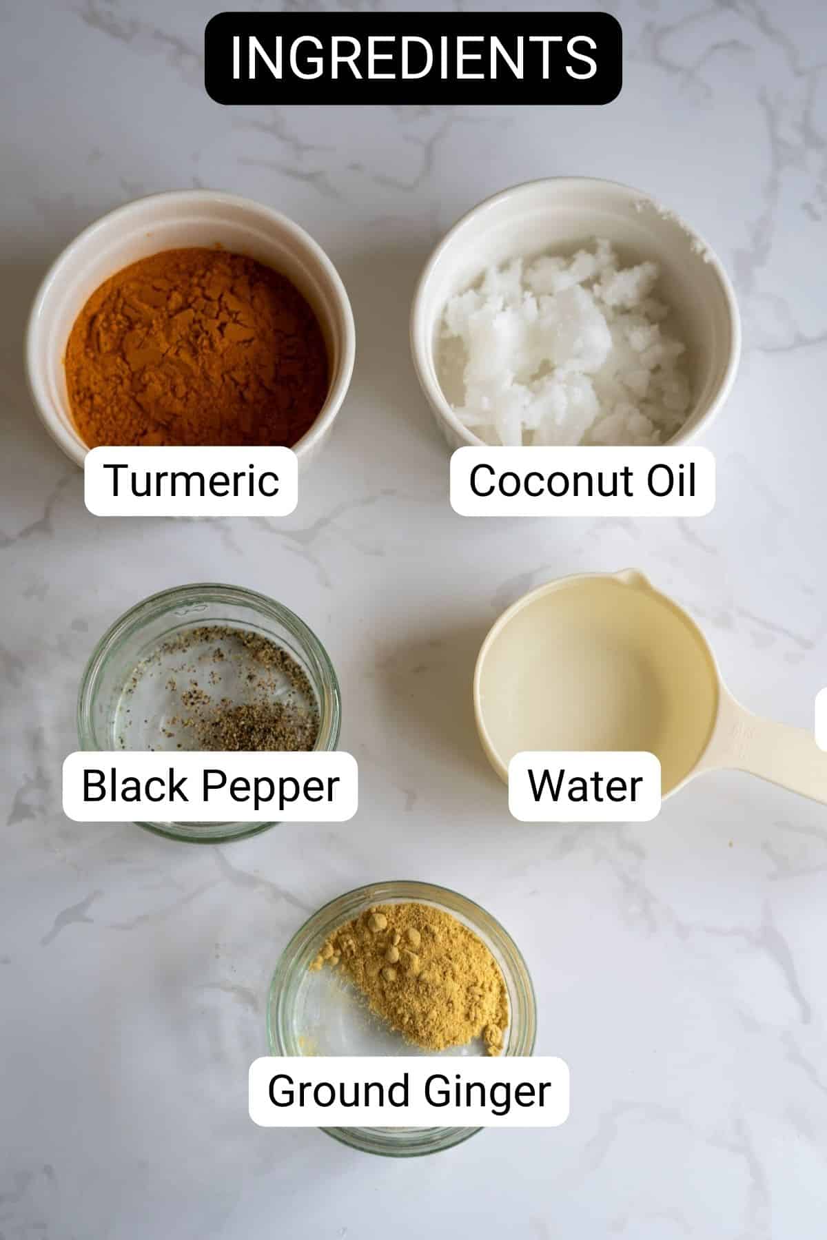Five labelled ingredients—turmeric, coconut oil, black pepper, water, and ground ginger—are arranged in bowls and a measuring jug on a white marble surface.