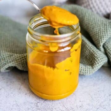 An open glass jar filled with thick yellow paste, with a spoon holding some of the paste above the jar, and a textured green cloth in the background.