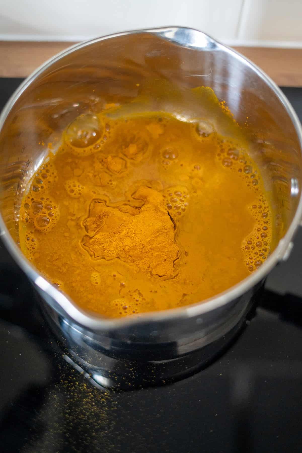 A stainless steel pot on a hob containing yellow liquid and turmeric powder.