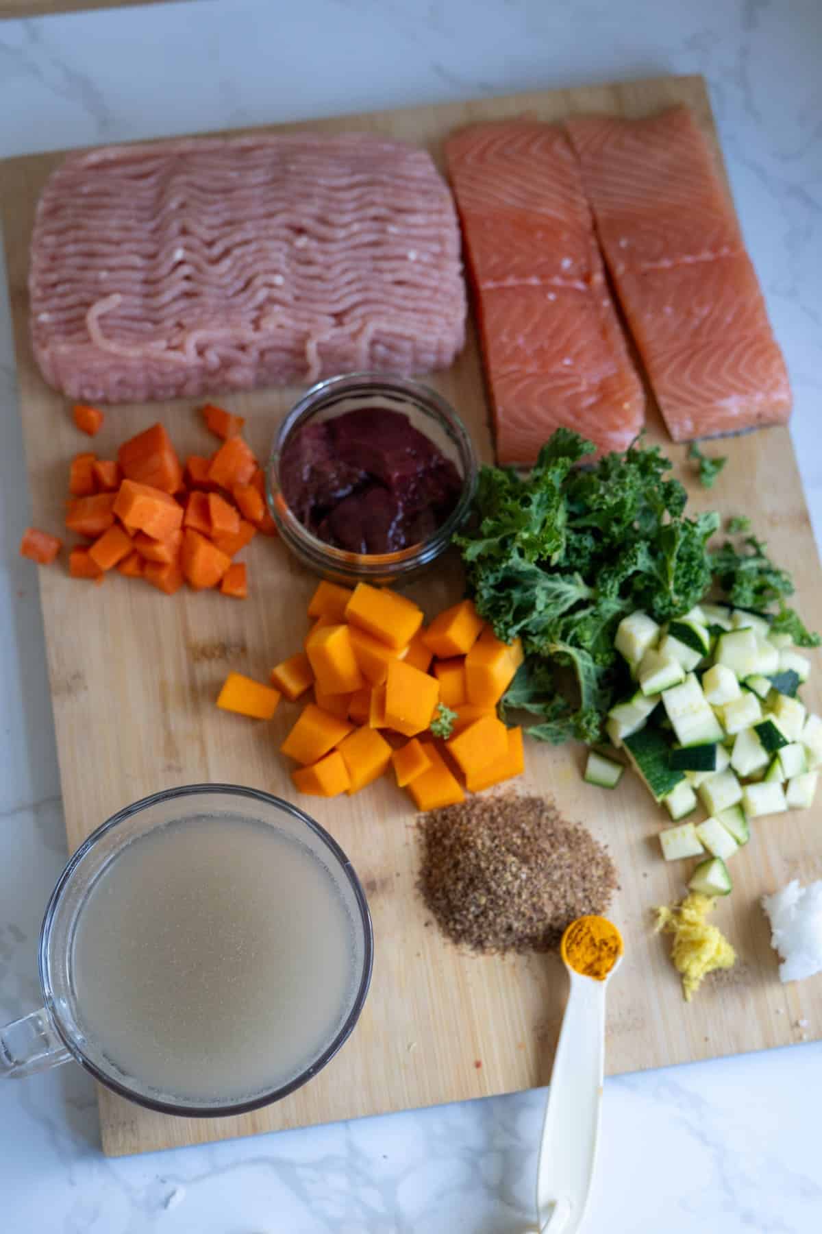 Chopped salmon, minced turkey and salmon dog food, carrots, squash, courgette, kale, linseed, liver, ginger, garlic, turmeric, and stock arranged on a wooden chopping board.