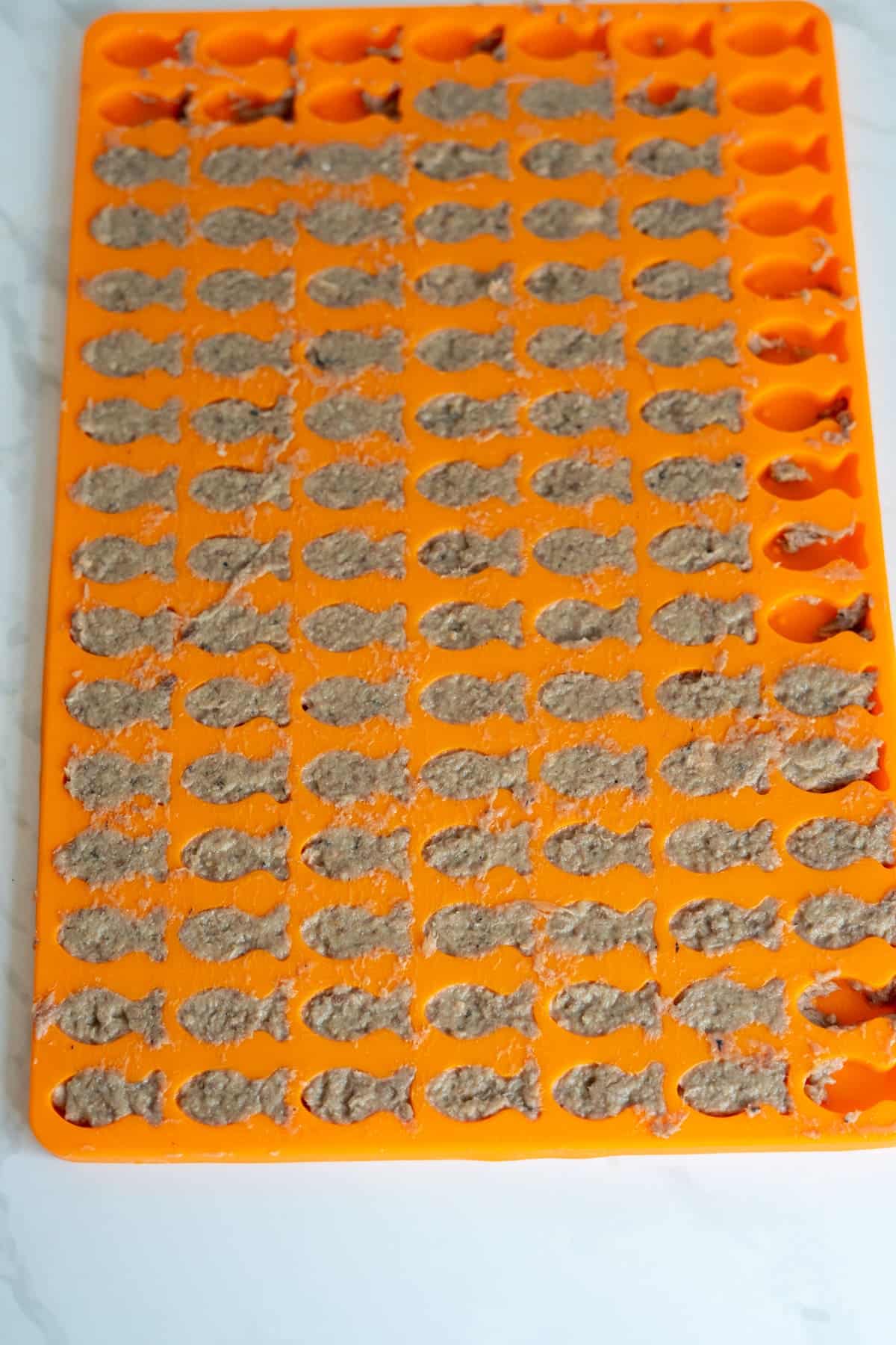 An orange silicone mould tray filled with a brown sardine cat treats mixture shaped like small fish, sitting on a white surface.