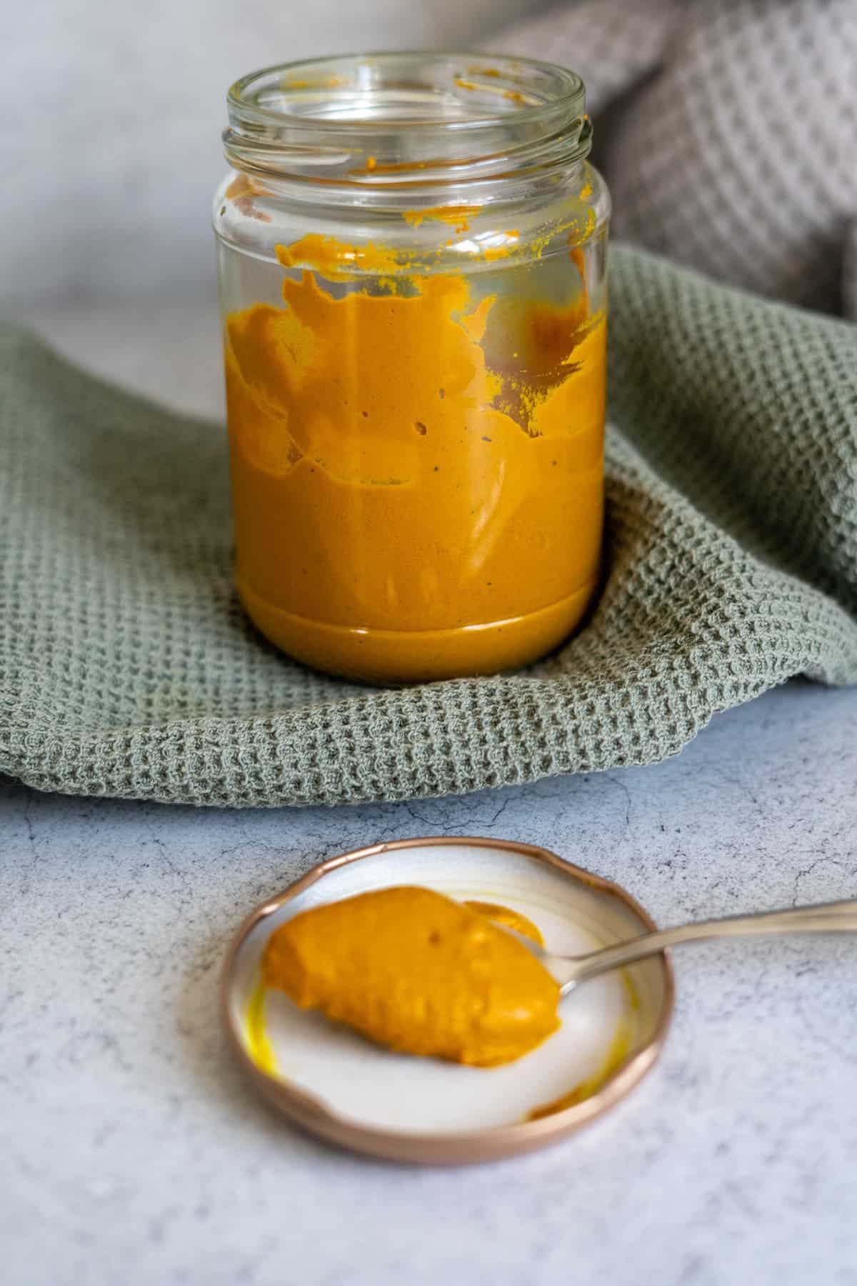 A jar of thick yellow-orange paste sits on a green textured cloth, with a spoonful of the paste on a small plate in front.