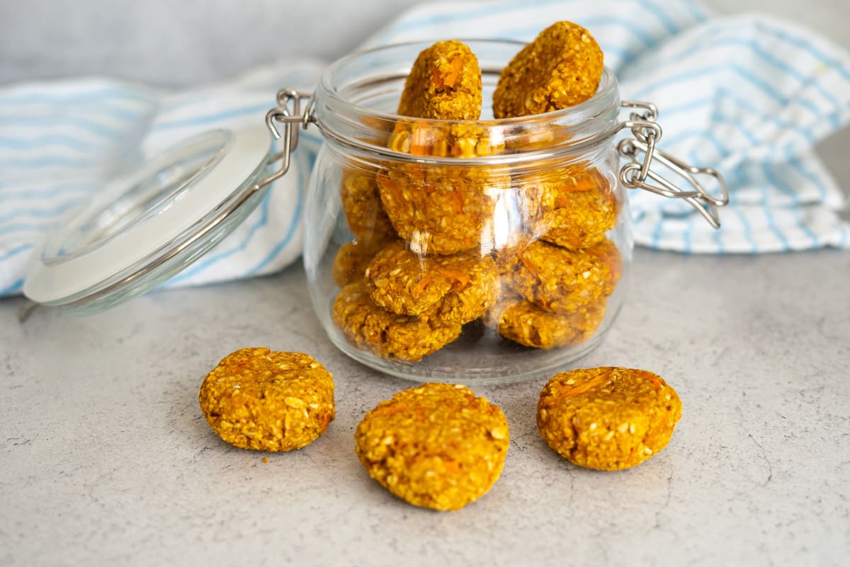 A glass jar filled with round, golden oat biscuits sits on a grey surface, with three biscuits placed in front of the jar. A blue and white striped cloth is in the background.