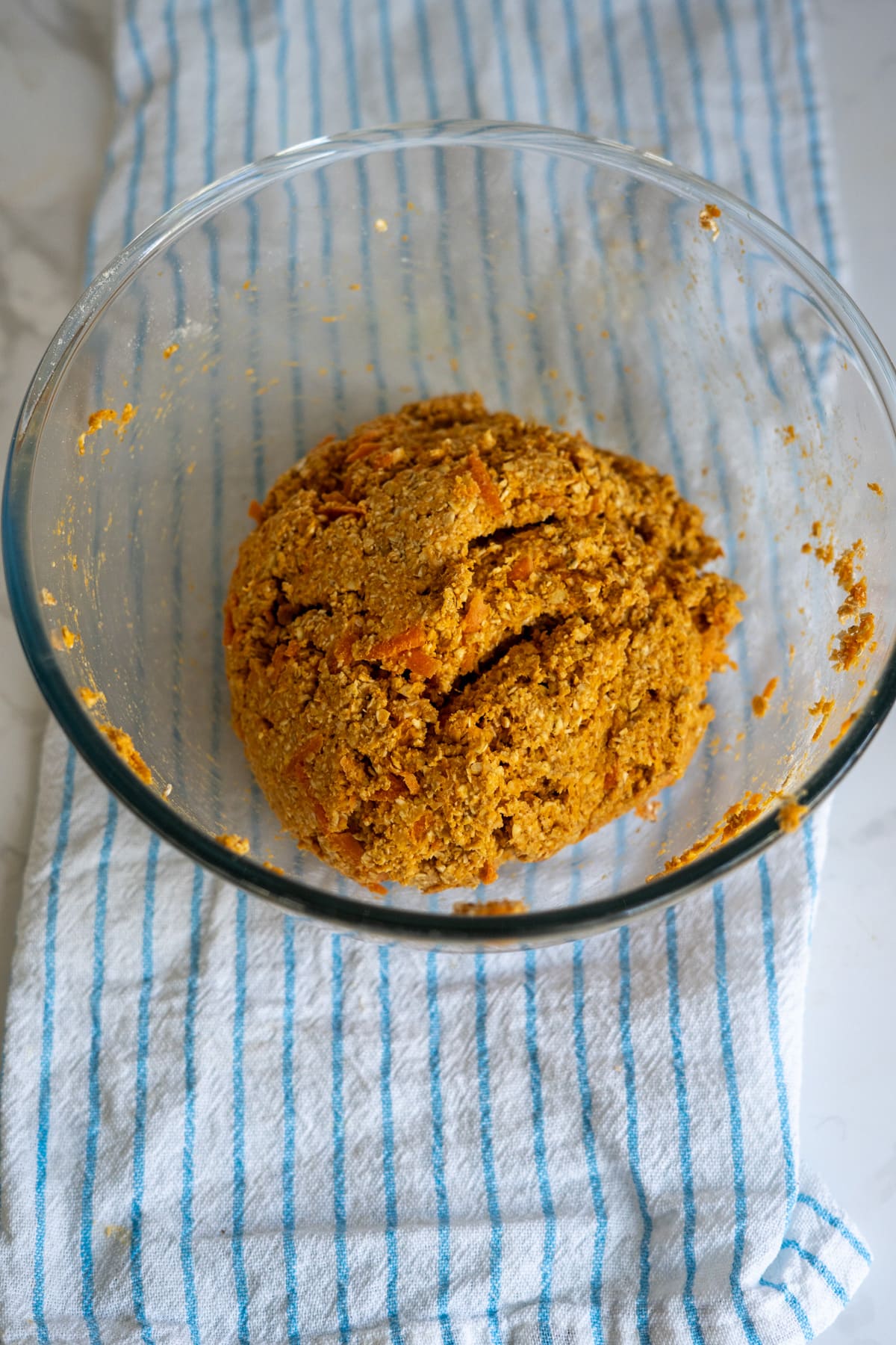 A glass bowl containing a ball of orange-coloured dough sits on a blue and white striped kitchen towel.