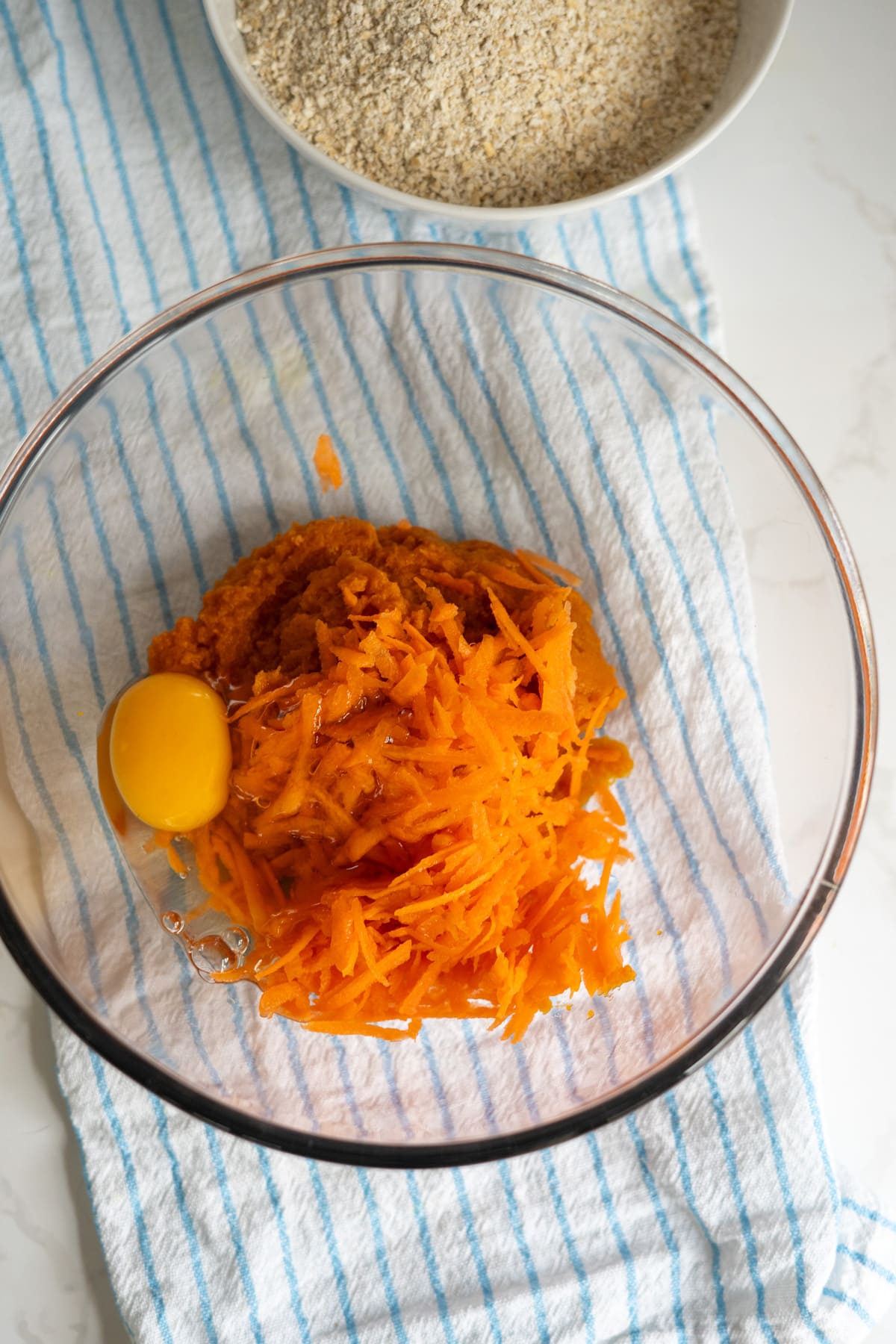 A glass bowl with grated carrots, an egg, and mashed sweet potato on a striped tea towel, with a bowl of breadcrumbs in the background.