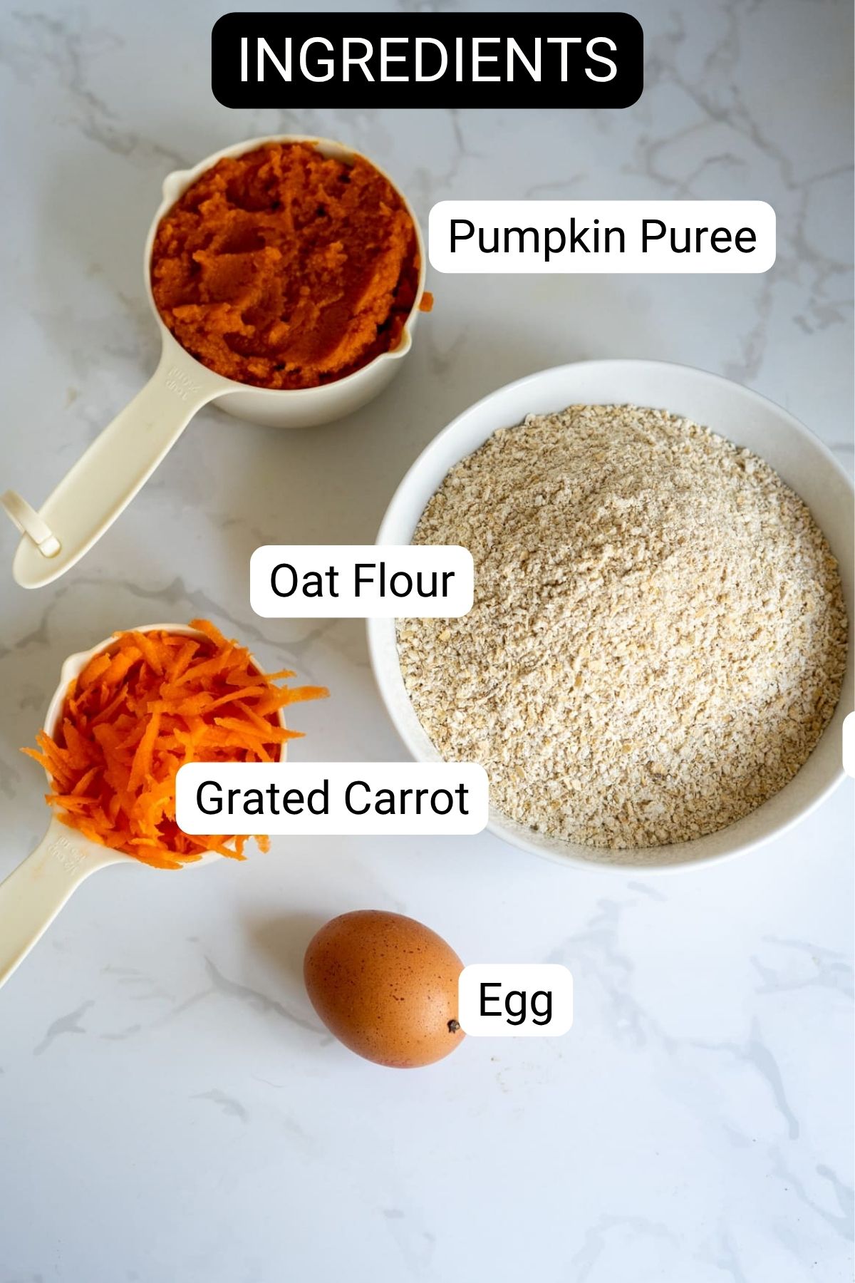 A flat lay of ingredients including pumpkin purée, oat flour, grated carrot, and an egg, each labelled, arranged on a white surface.