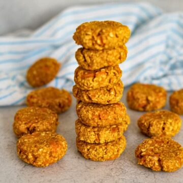 A stack of eight homemade oat biscuits sits on a light surface, with more biscuits scattered around and a blue-striped cloth in the background.