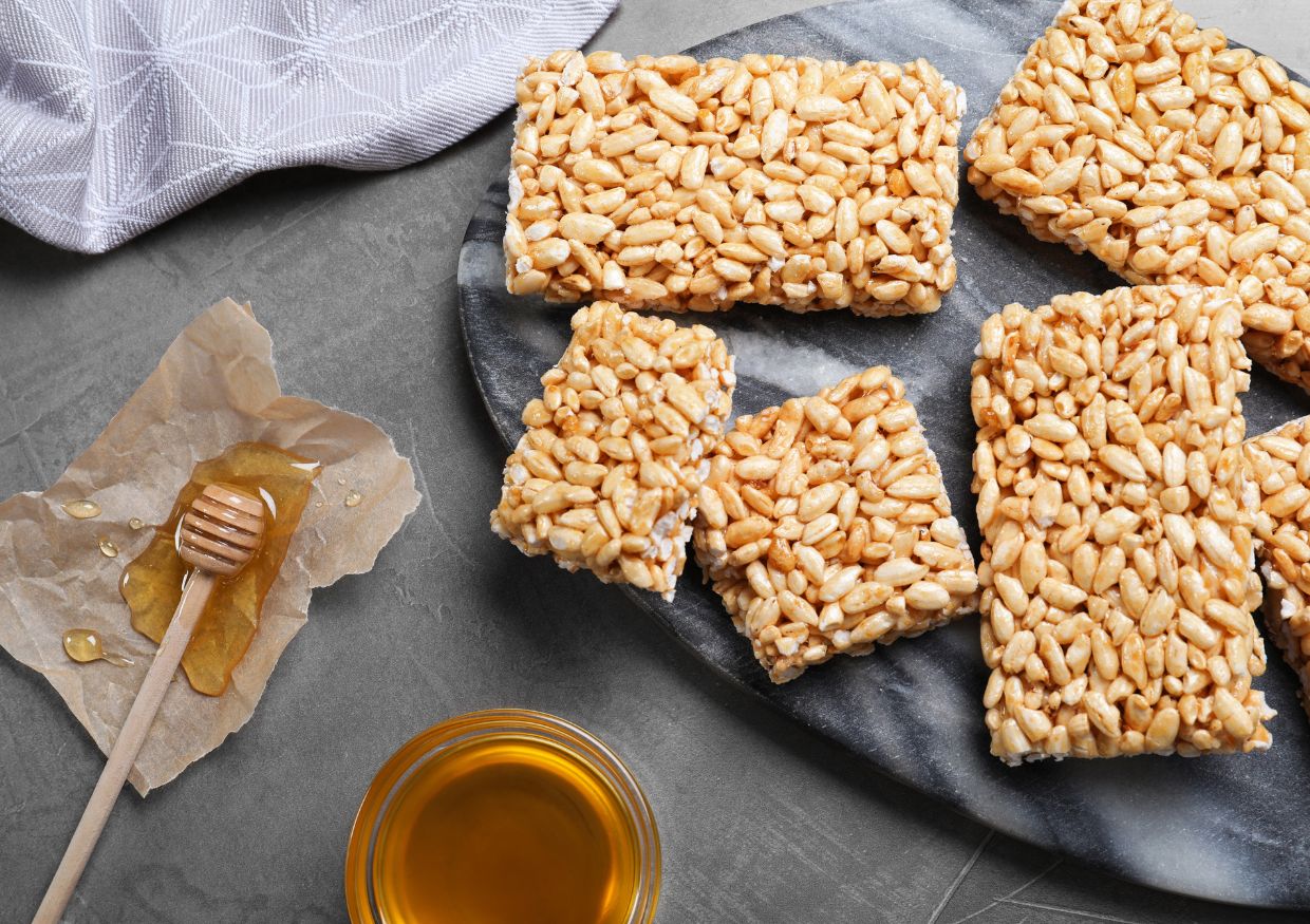 Rectangular puffed rice bars on a dark platter with a honey dipper, honey, and a serviette on a grey surface.
