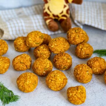 A group of round, orange veg balls arranged on a light surface with a chequered cloth, a soft toy, and some fresh dill in the background.