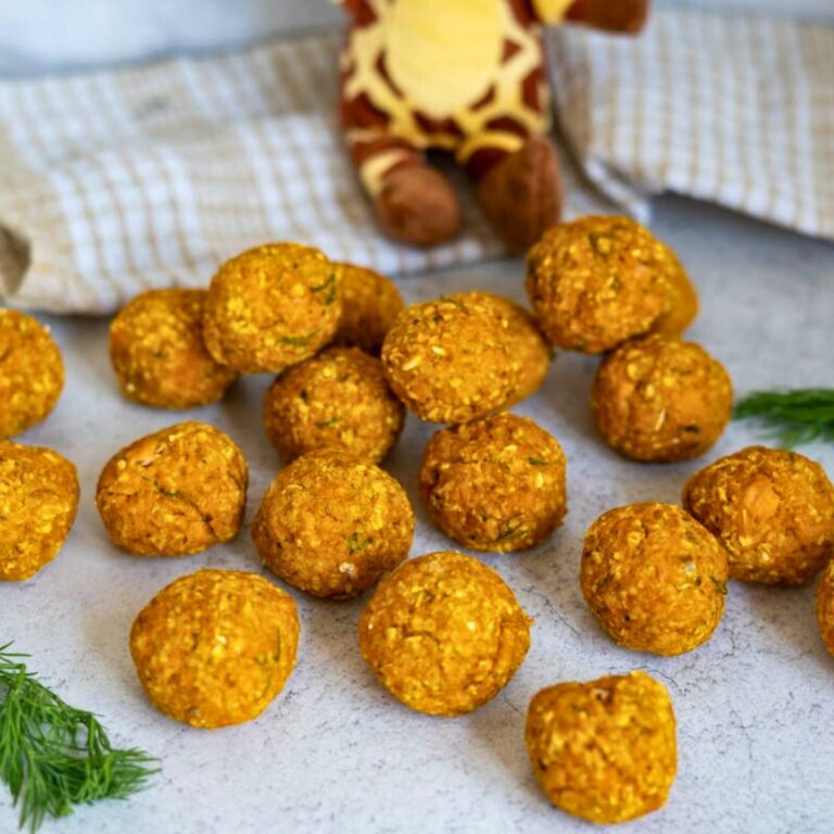 A group of round, orange veg balls arranged on a light surface with a chequered cloth, a soft toy, and some fresh dill in the background.