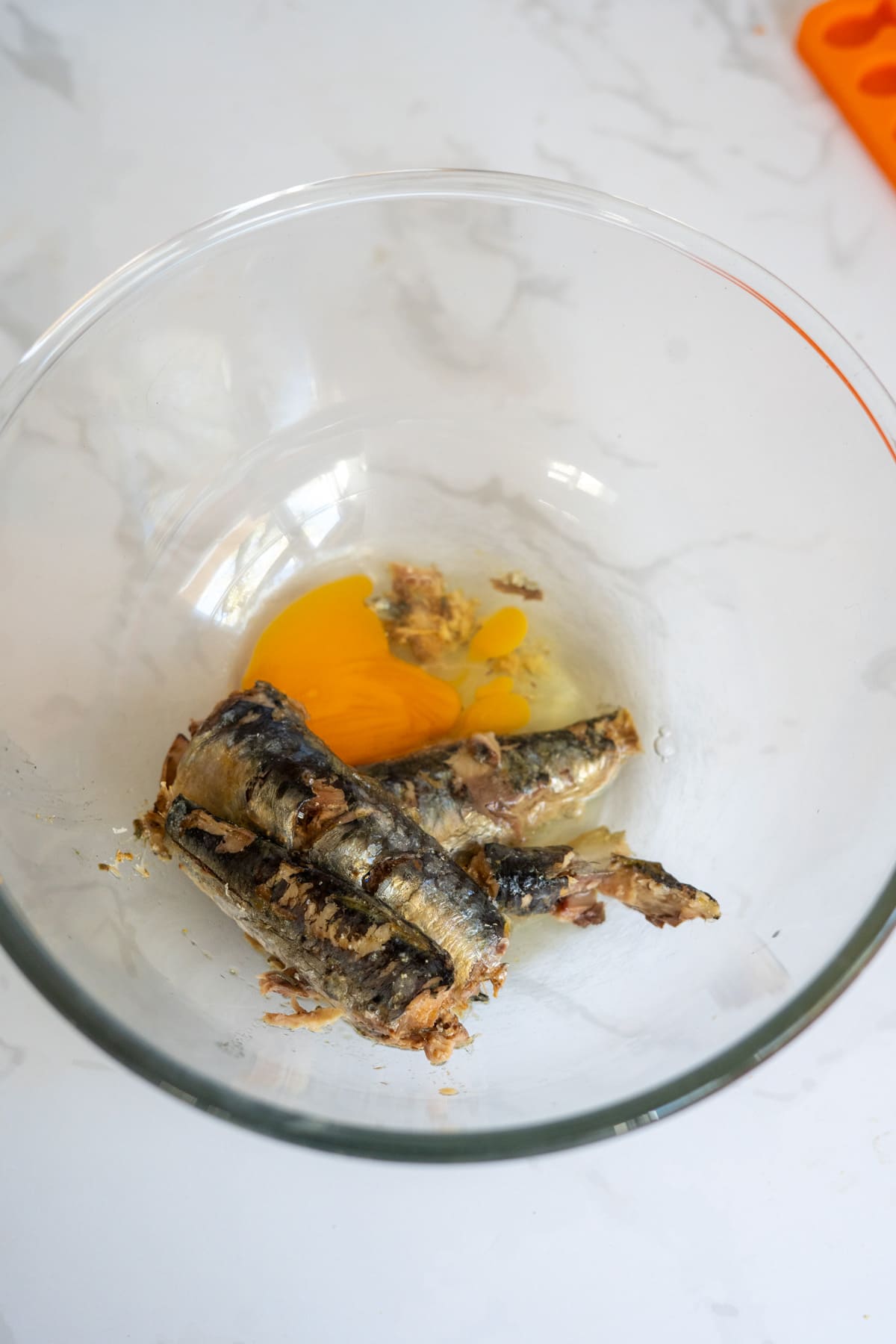 A clear glass bowl containing two raw eggs and three tinned sardines, perfect for making homemade sardine cat treats, sits on a white marble surface.