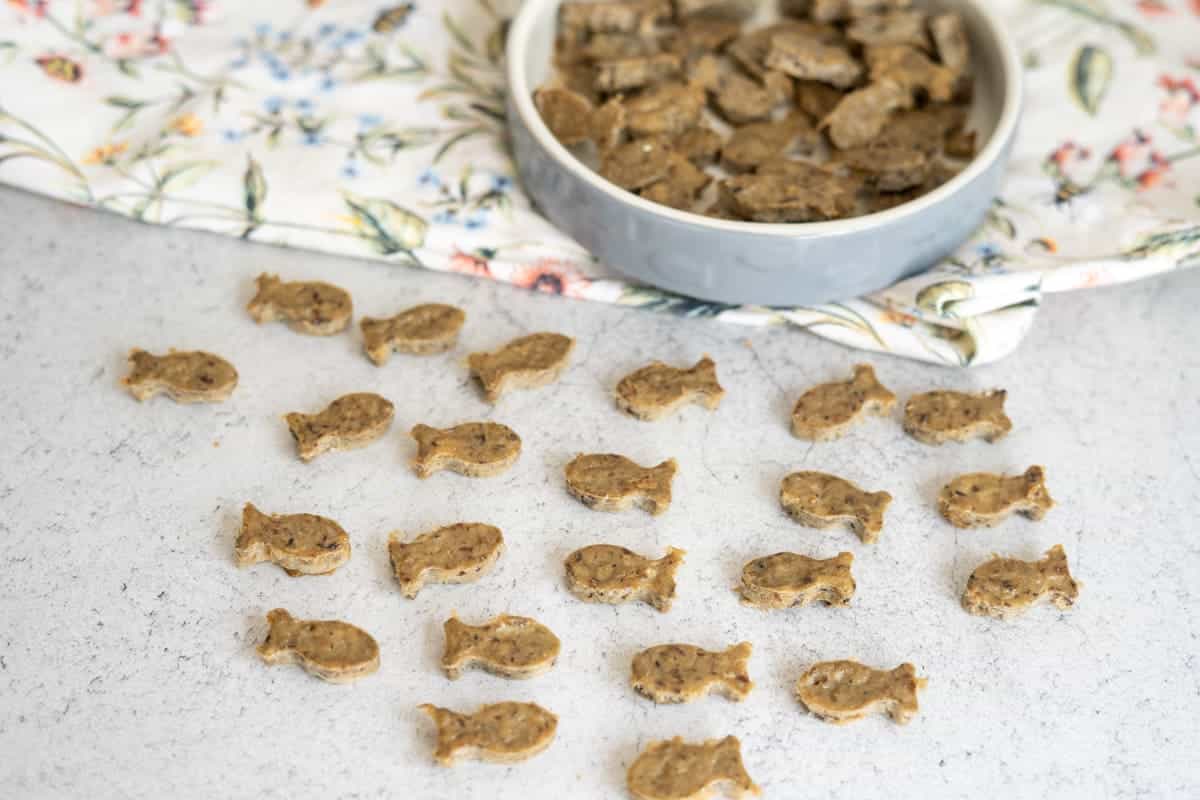 Small, fish-shaped biscuits are arranged in rows on a light surface, with more biscuits in a round dish placed on a floral cloth in the background.