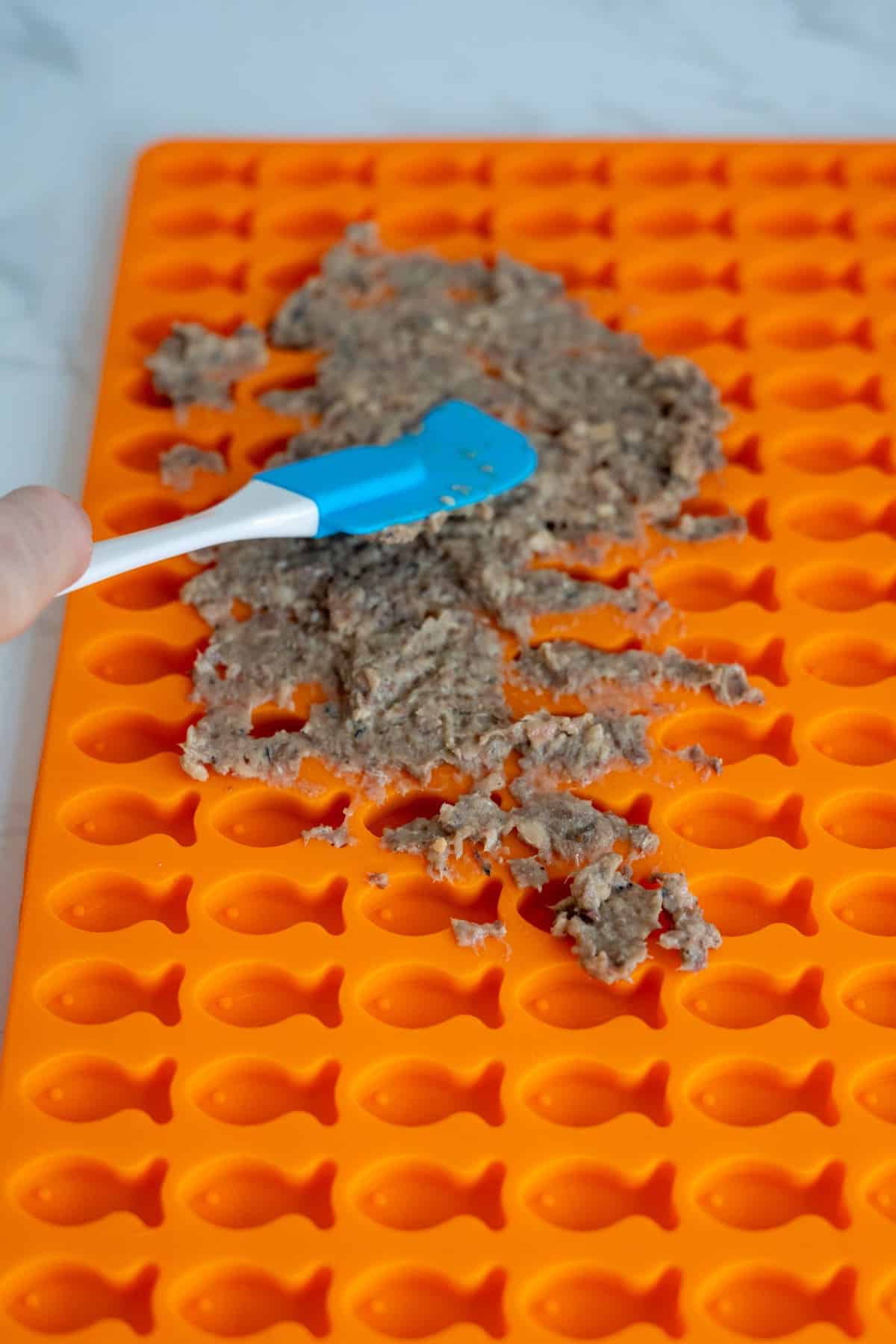A hand uses a blue spatula to spread a greyish sardine cat treats mixture onto an orange silicone mould with fish-shaped cavities.