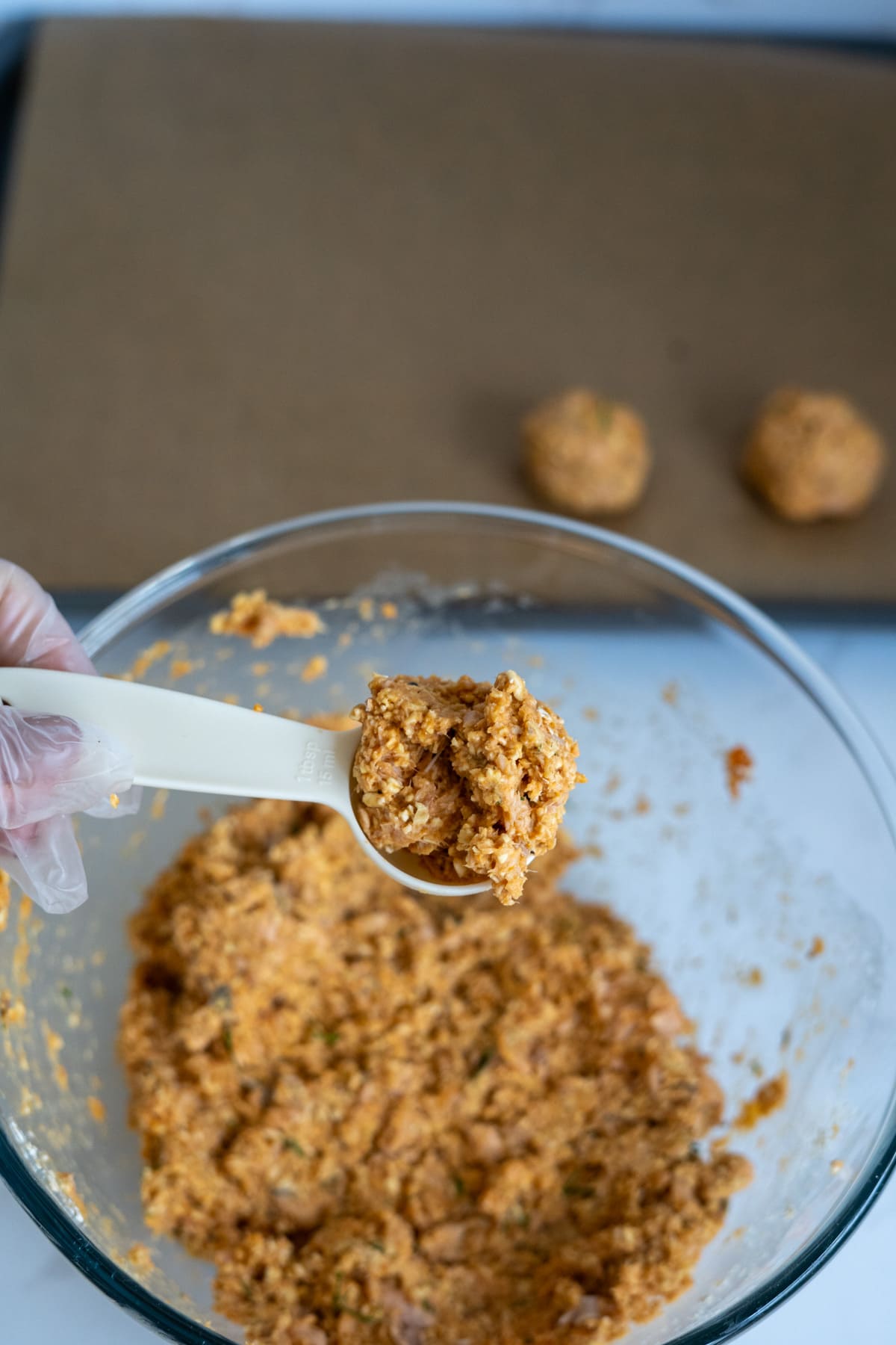 A gloved hand holds a measuring spoon of orange mixture over a glass bowl, with two shaped mounds of salmon meatballs for dogs on a tray in the background.