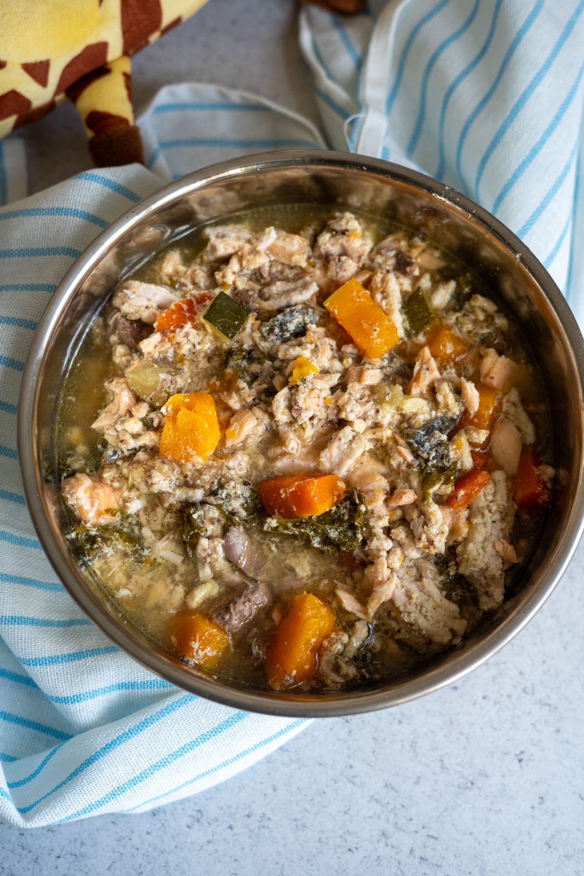A metal bowl filled with a chunky mixture of turkey and salmon dog food, cooked meat, carrots, courgette, leafy greens, and broth, placed on a blue and white striped cloth.