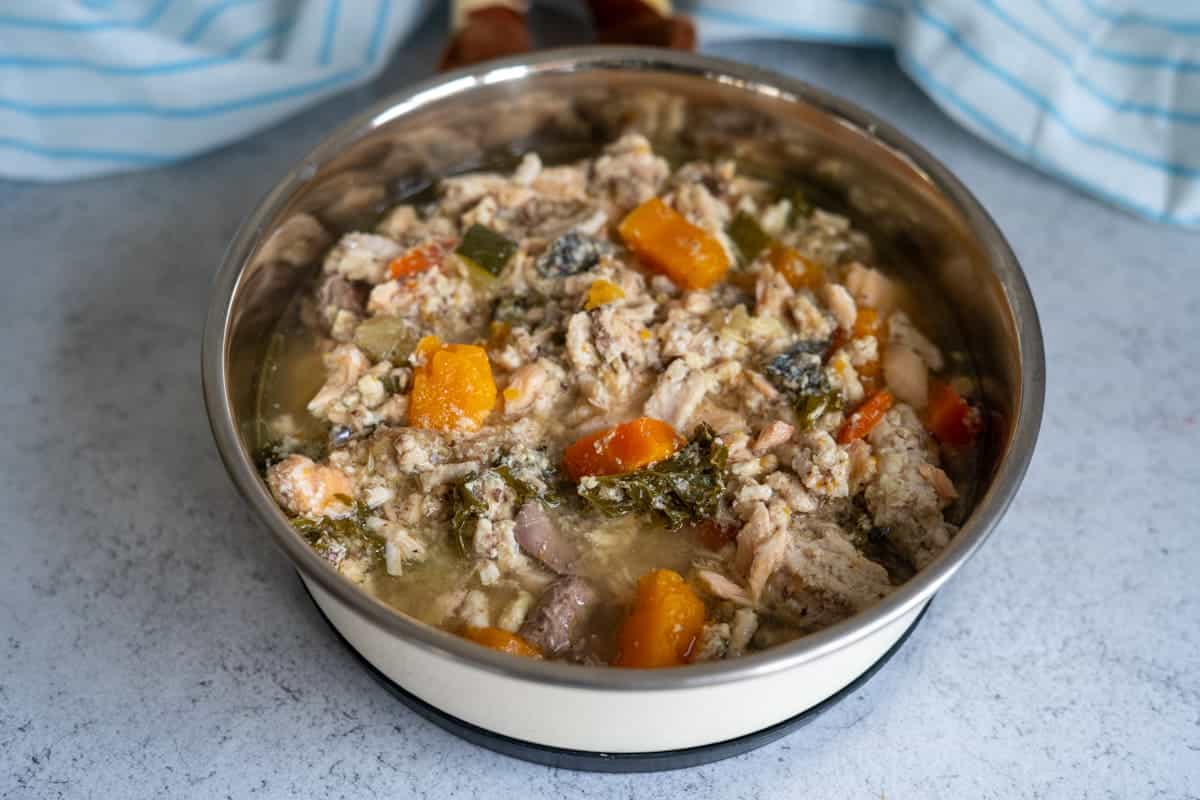 A metal bowl filled with homemade dog food containing chunks of meat, carrots, courgette, kale, and other vegetables in broth.