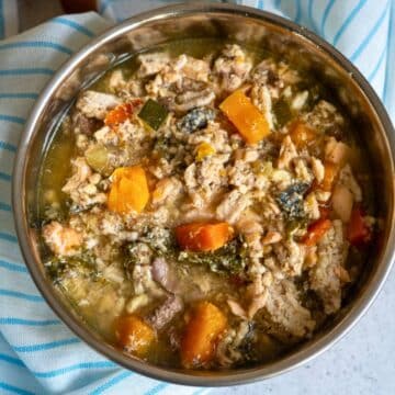 A stainless steel bowl filled with a chunky mixture of cooked meat, carrots, and leafy greens in a broth, placed on a blue and white striped cloth.