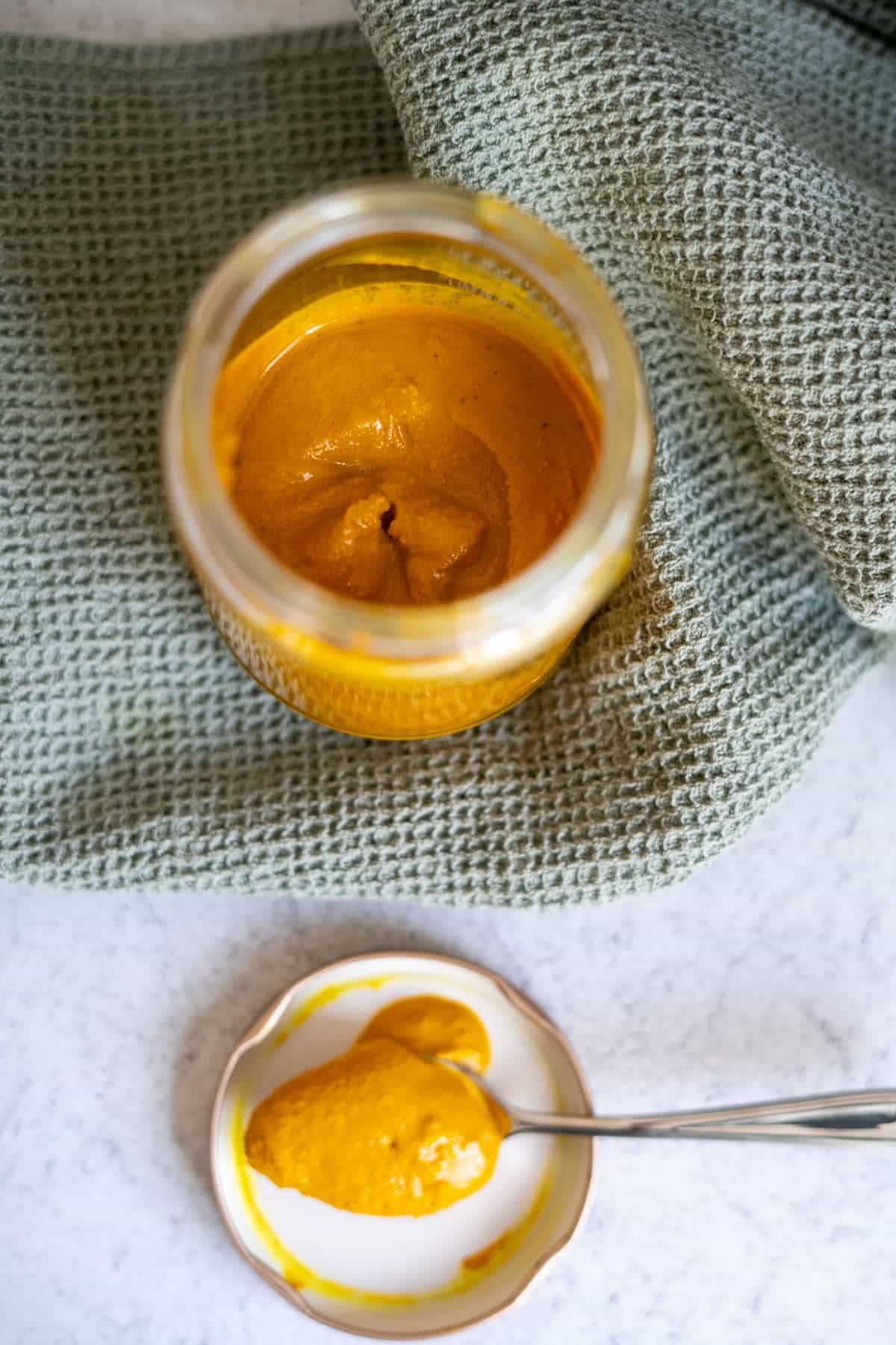 Open jar of yellow mustard on a green textured cloth, with a spoonful of mustard on a small plate beside it.