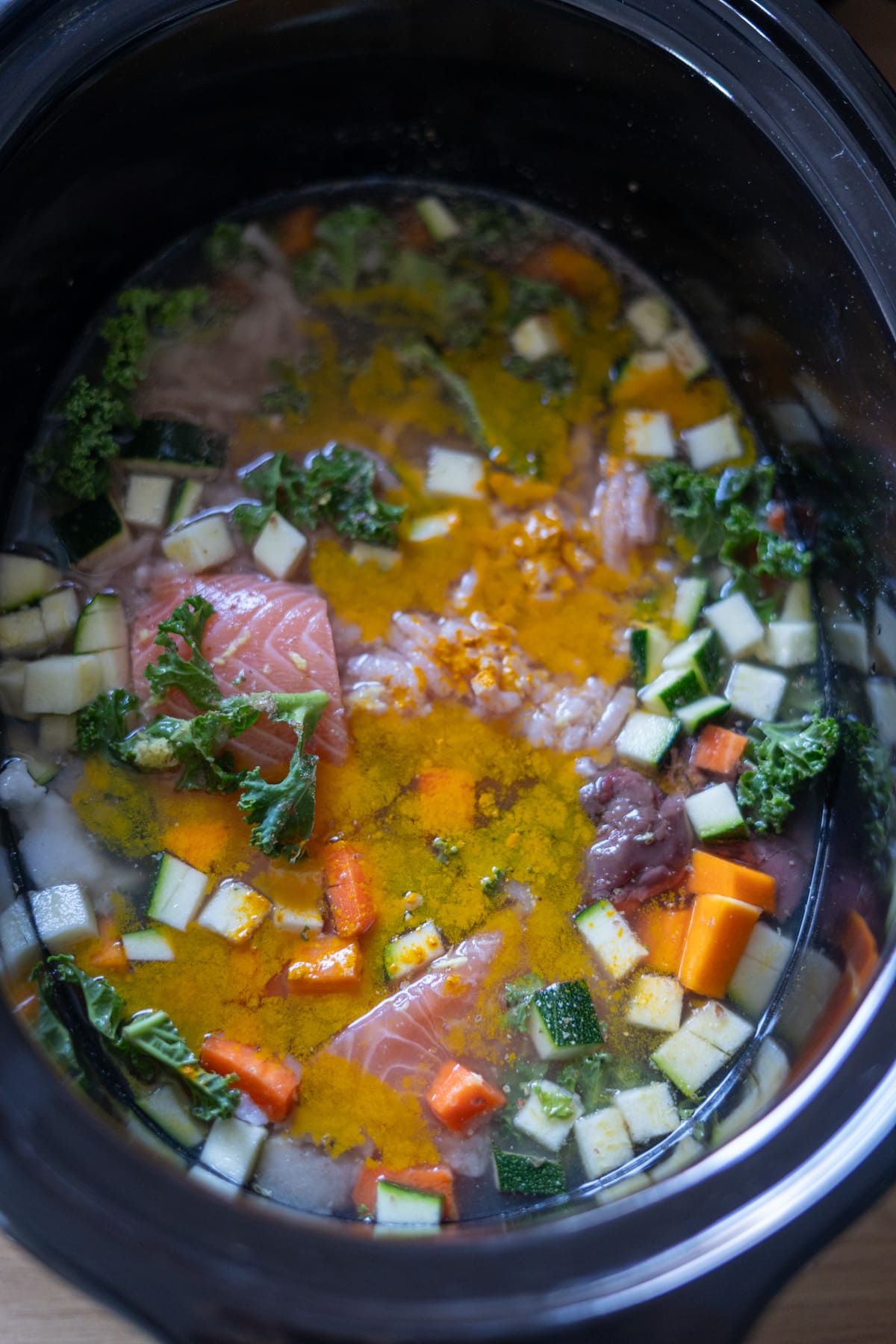 Chunks of raw salmon, chopped courgette, carrots, kale, and spices in broth inside a slow cooker—perfect for making nutritious turkey and salmon dog food your pup will love.