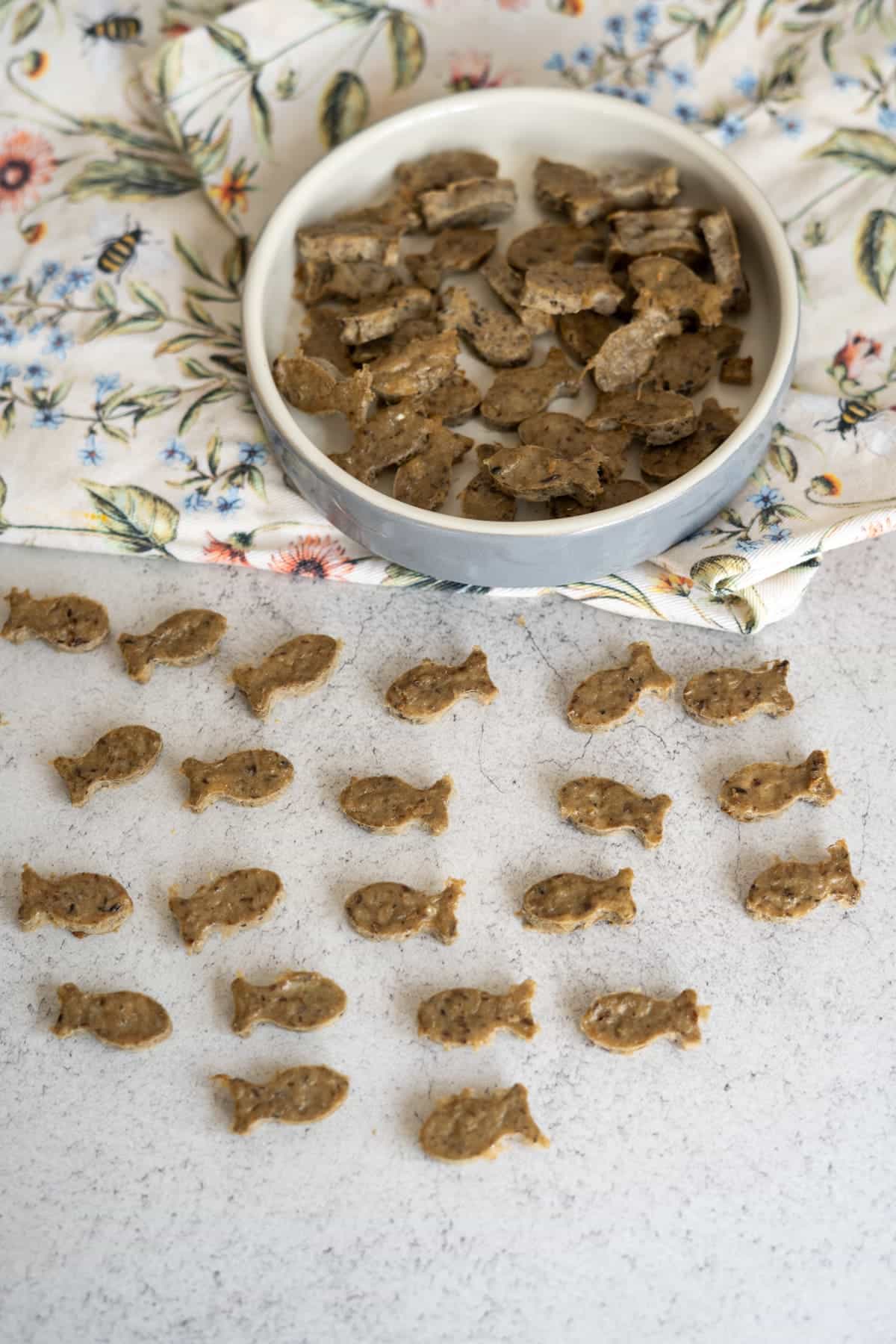A round dish filled with small, fish-shaped sardine cat treats sits on a floral cloth, with more treats arranged in rows on a light worktop.