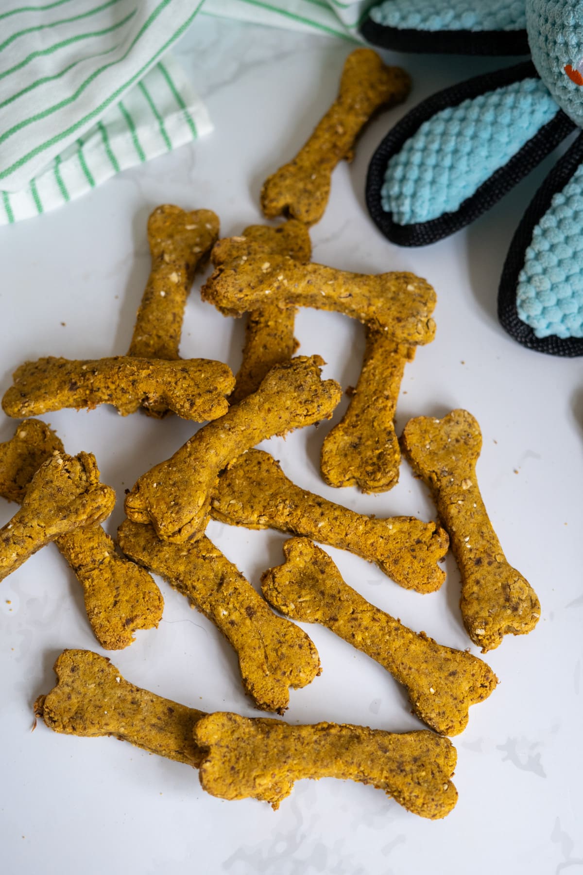 Lamb liver dog treats, bone-shaped and tasty, are scattered on a white surface next to a blue soft toy and a green-striped cloth.
