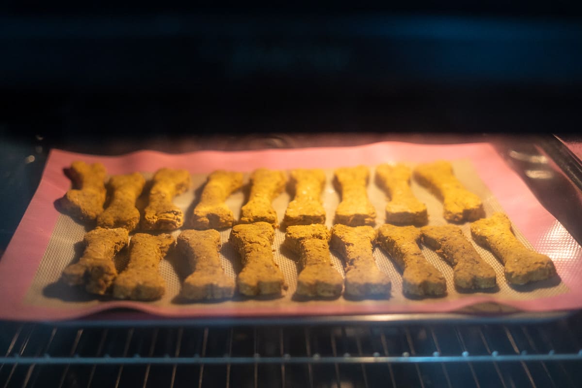 Rows of bone-shaped dog biscuits baking on a silicone mat inside an oven.