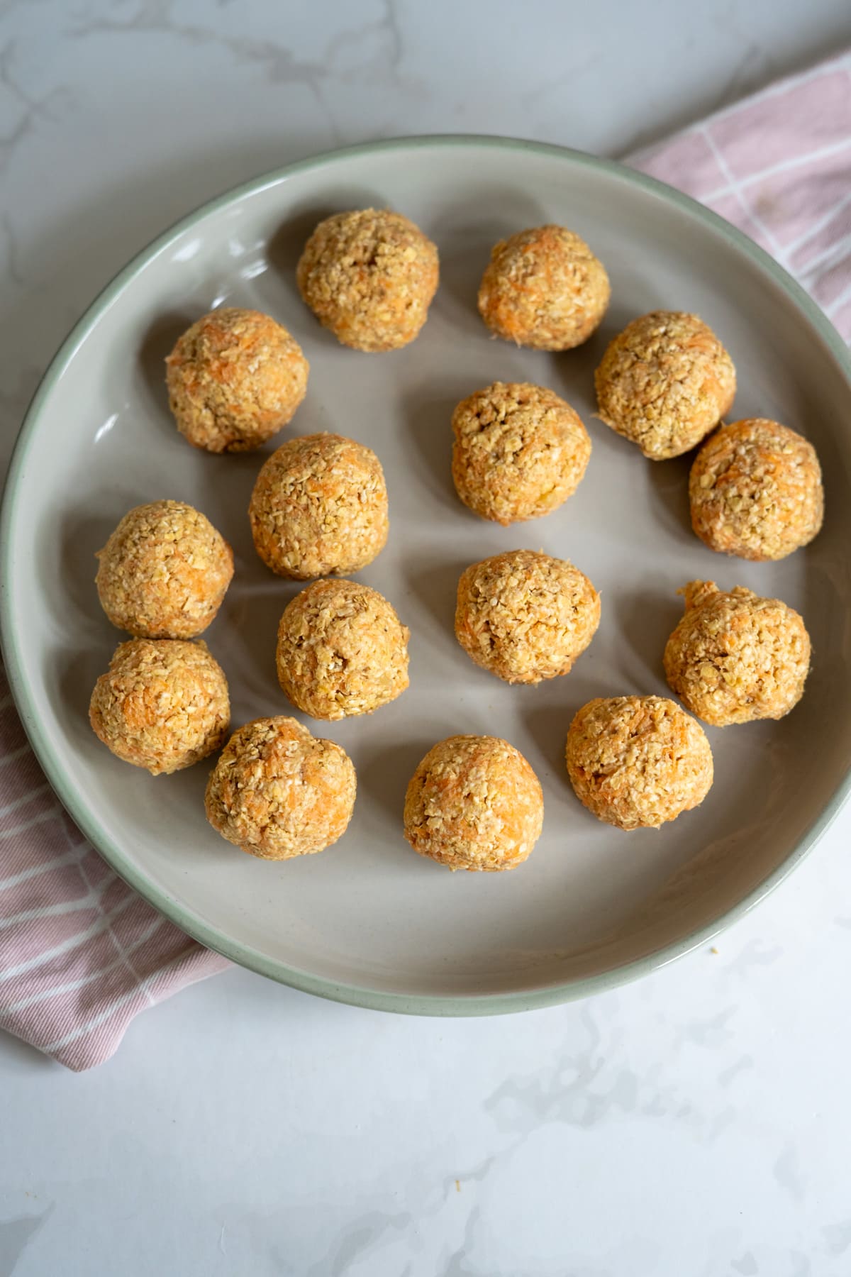 A round plate with fifteen banana carrot dog treats shaped into balls, arranged in a circular pattern, placed on a light pink cloth on a marble surface.