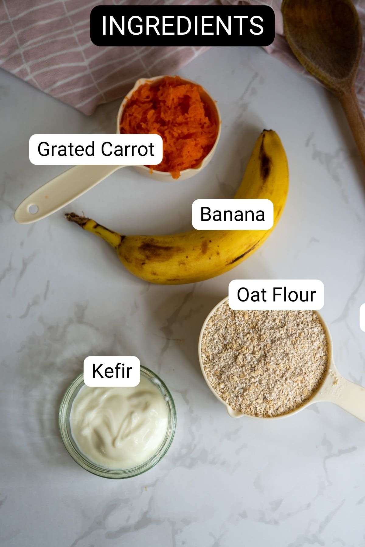 Four labelled ingredients—grated carrot, banana, oat flour, and kefir—are arranged on a marble surface with a wooden spoon and a cloth visible in the background.