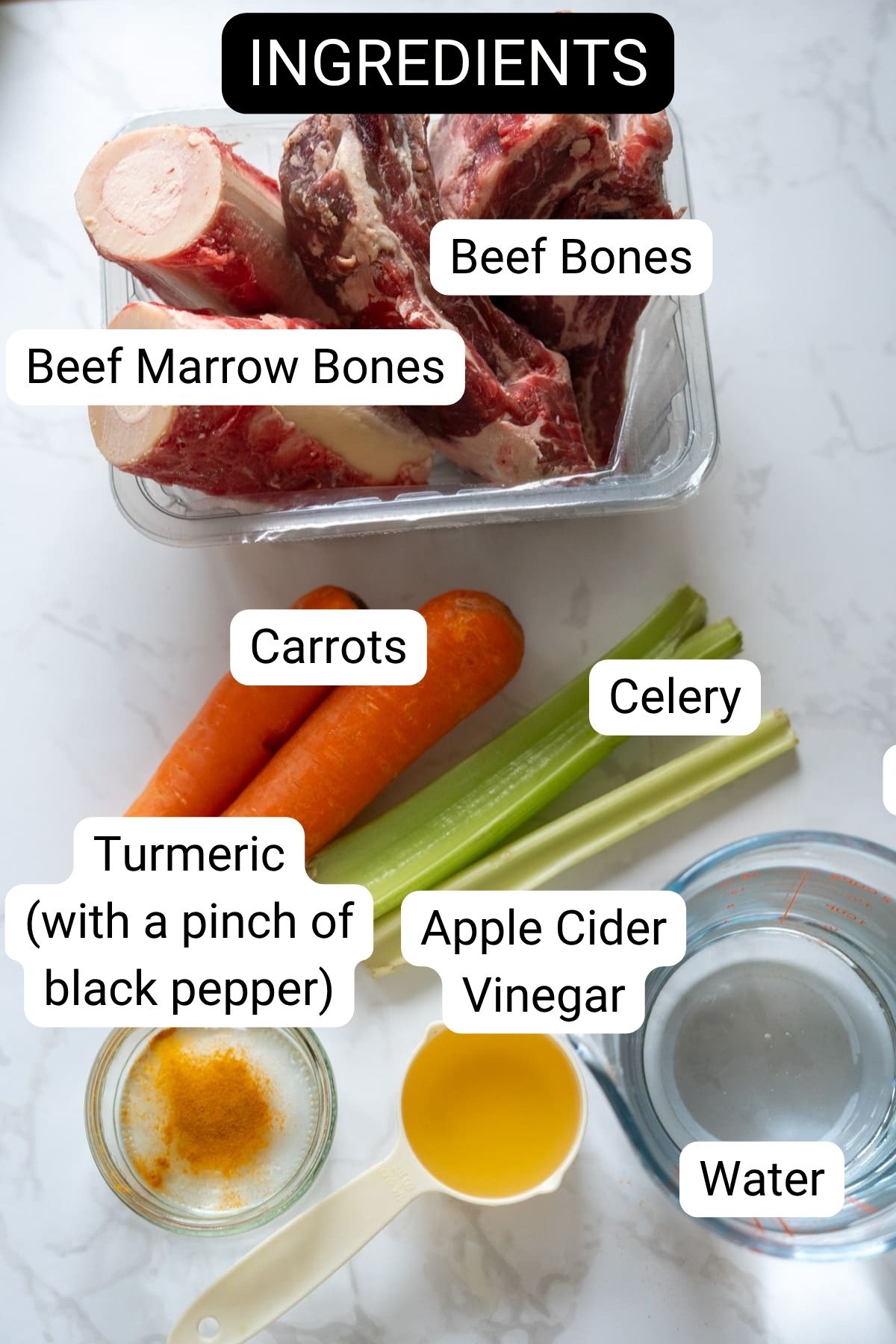 A selection of ingredients for beef bone broth for dogs: beef bones, marrow bones, carrots, celery, turmeric with black pepper, apple cider vinegar, and a bowl of water on a white surface.