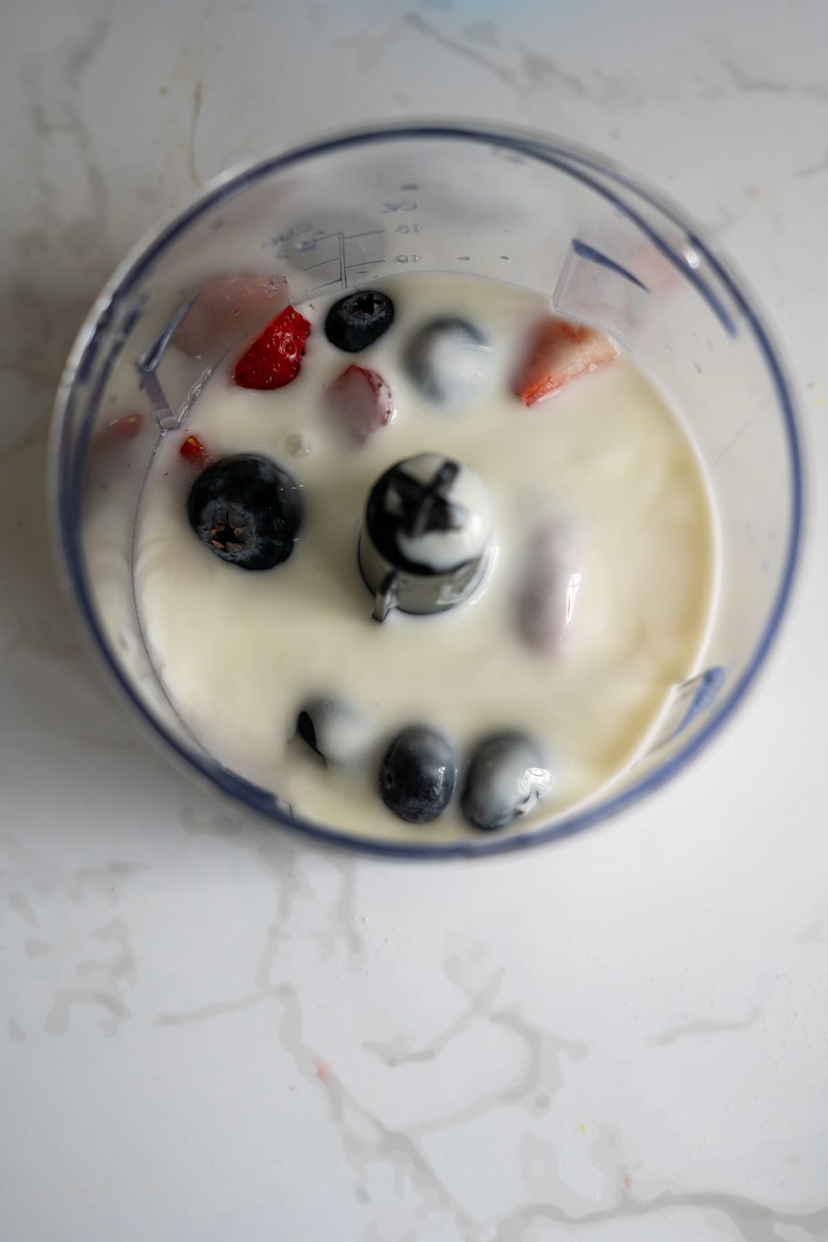 A blender beaker filled with milk, blueberries, strawberries, and ice cubes, viewed from above on a white worktop.