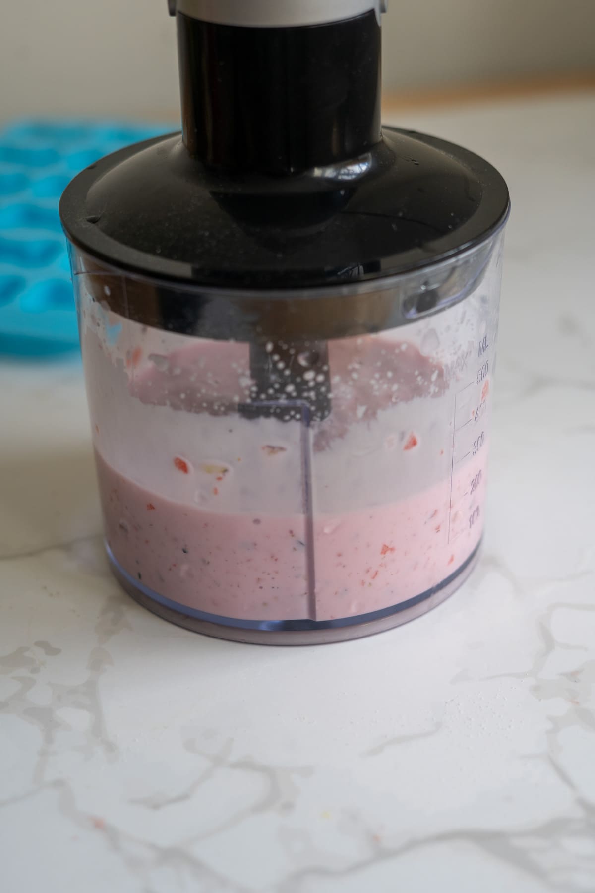 Clear food processor bowl containing a pink mixture sits on a white worktop with a blue silicone mould in the background.
