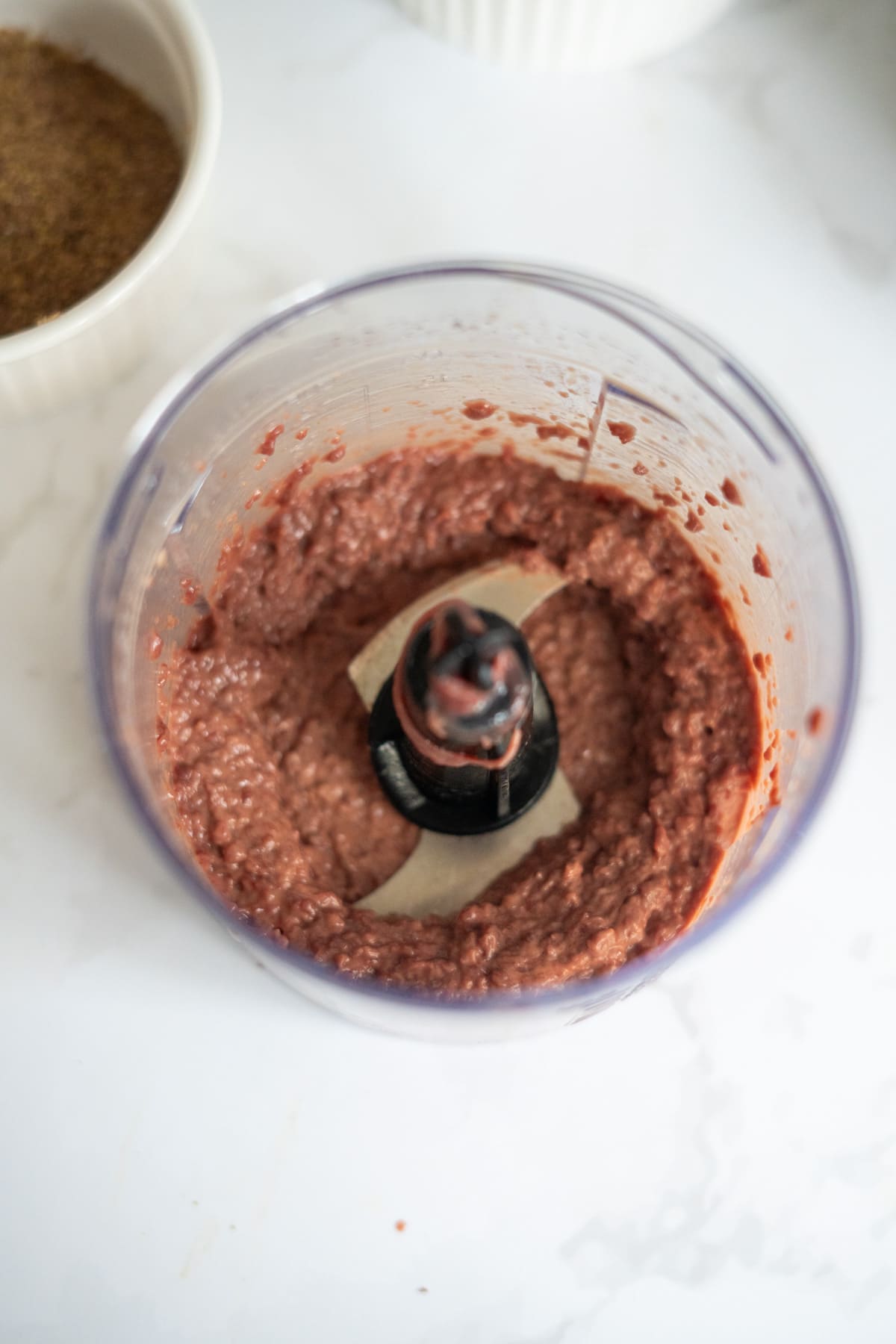 A food processor bowl containing blended reddish-brown mixture sits on a white surface, with part of another bowl visible nearby.