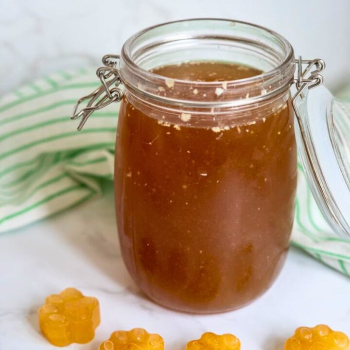 A glass jar filled with brown liquid, possibly honey or syrup, sits on a white surface beside a green-striped cloth and small yellow honeycomb-shaped sweets.