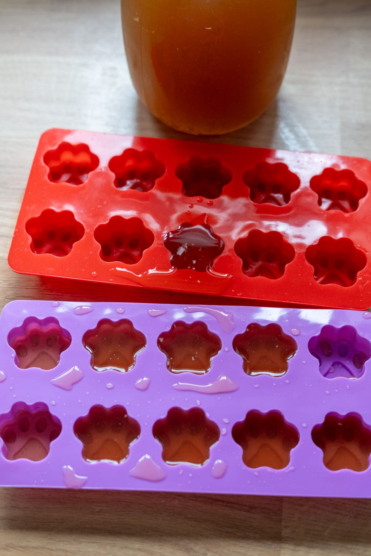 Two silicone paw-print moulds, one red and one purple, are filled with liquid on a wooden surface—perfect for making beef bone broth for dogs—with a jar of amber-coloured broth in the background.