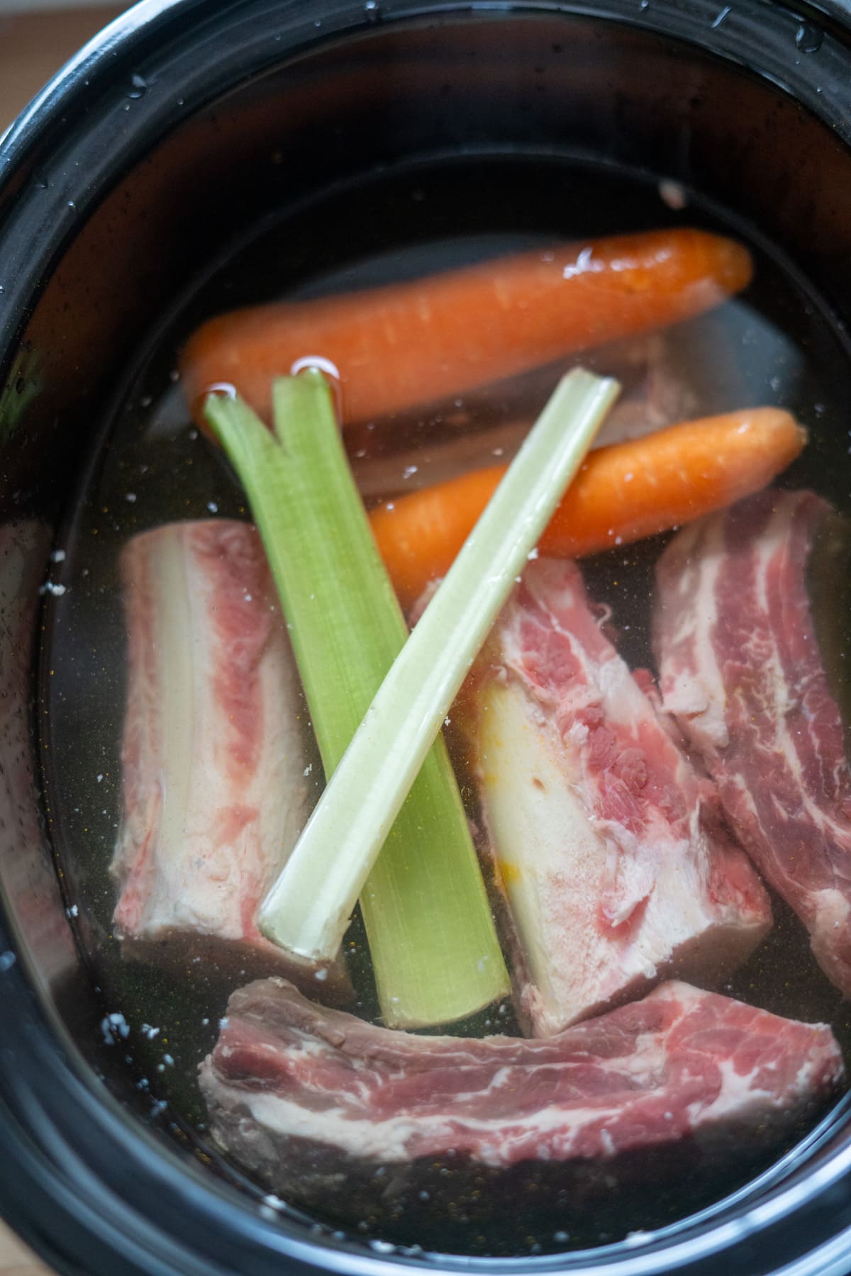 Raw beef ribs, whole carrots, and celery sticks submerged in water inside a slow cooker—perfect for making nutritious beef bone broth for dogs.