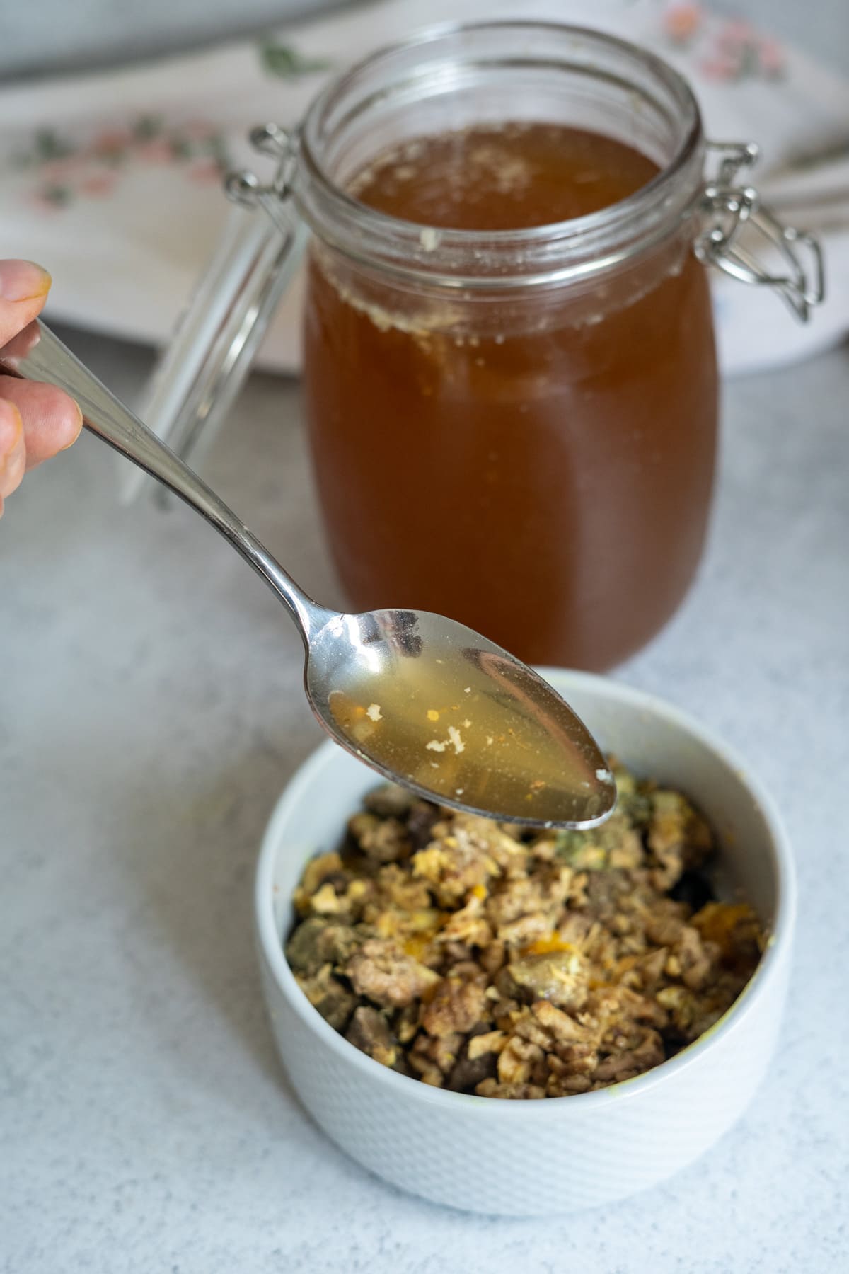 A hand holds a spoon of beef bone broth for dogs above a bowl of cooked minced meat and eggs, with a jar of broth in the background on a light worktop.