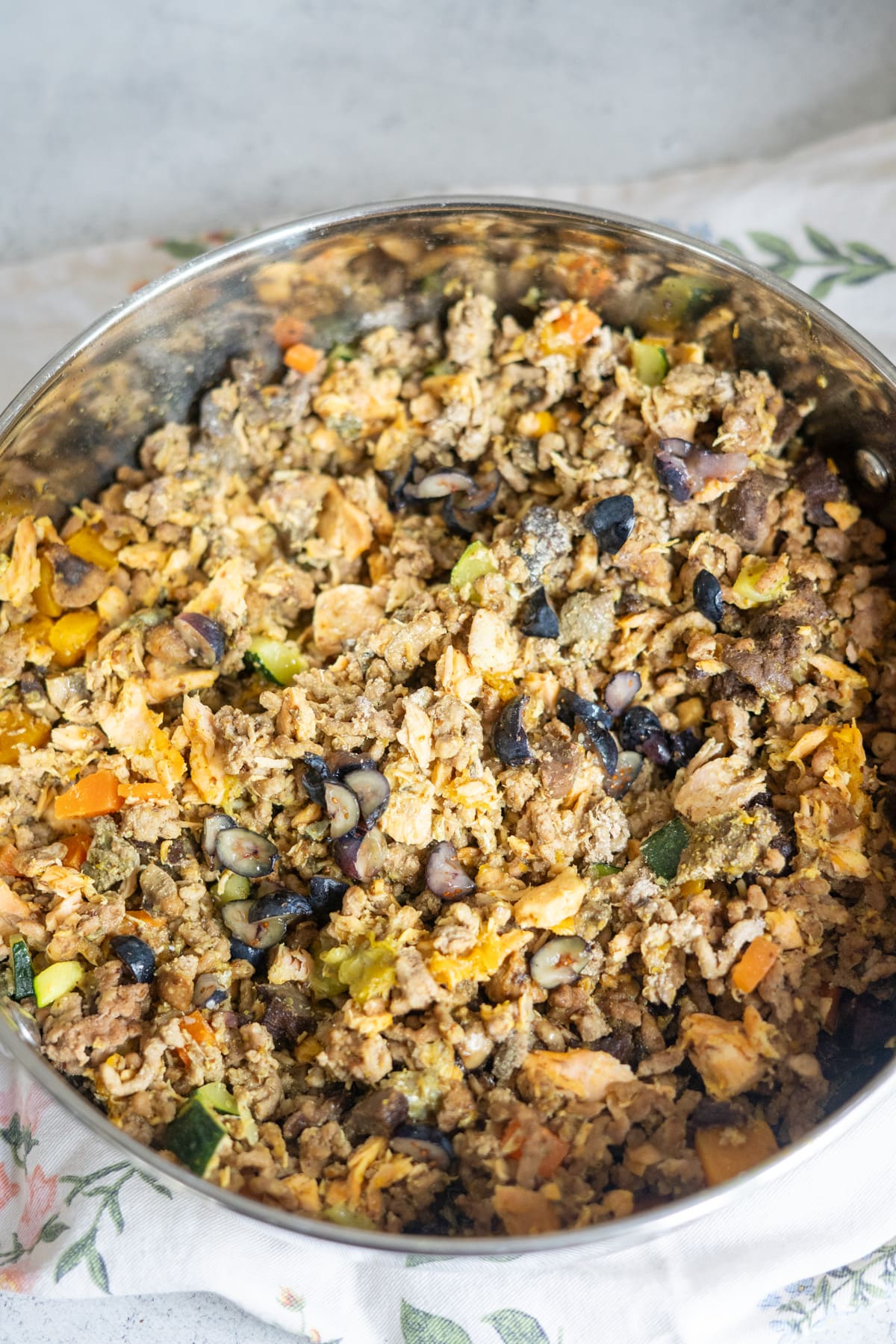 A close-up of a large metal bowl filled with a mixed blend of cooked minced meat, vegetables, and sliced black olives.
