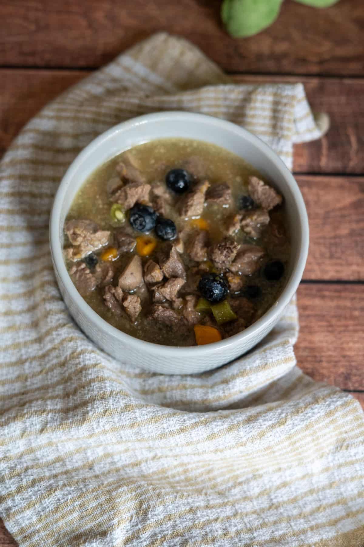 A bowl of lamb stew for dogs with chunks of meat, blueberries, and diced yellow vegetables sits on a striped cloth atop a wooden surface.