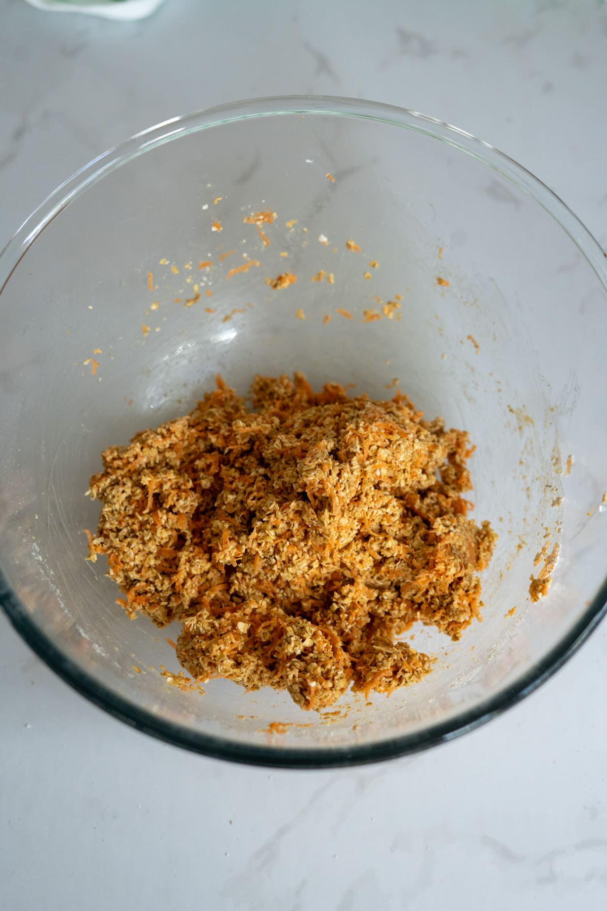 A glass bowl containing a mixture of grated carrots and oats for homemade duck dog treats sits on a white worktop.