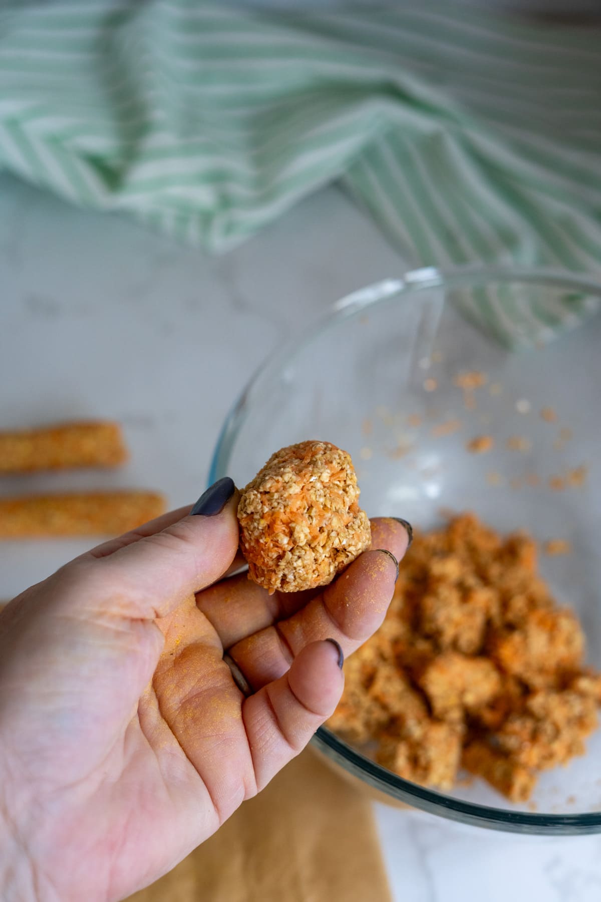 A hand holds a homemade duck dog treat over a glass bowl filled with similar nuggets, with a green striped cloth and breadsticks in the background.