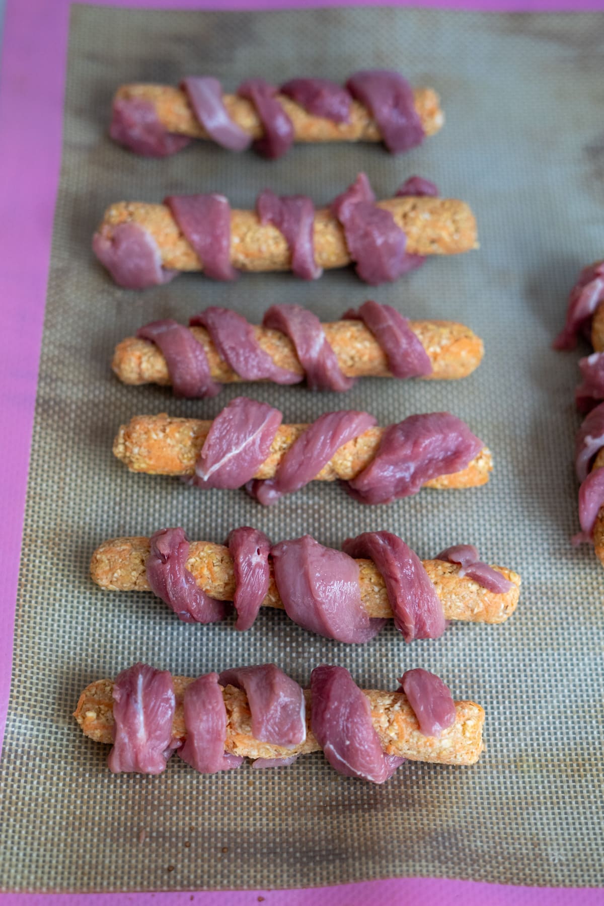 Homemade duck dog treats are made by wrapping raw meat around biscuit-like sticks and arranging them in rows on a silicone baking mat.