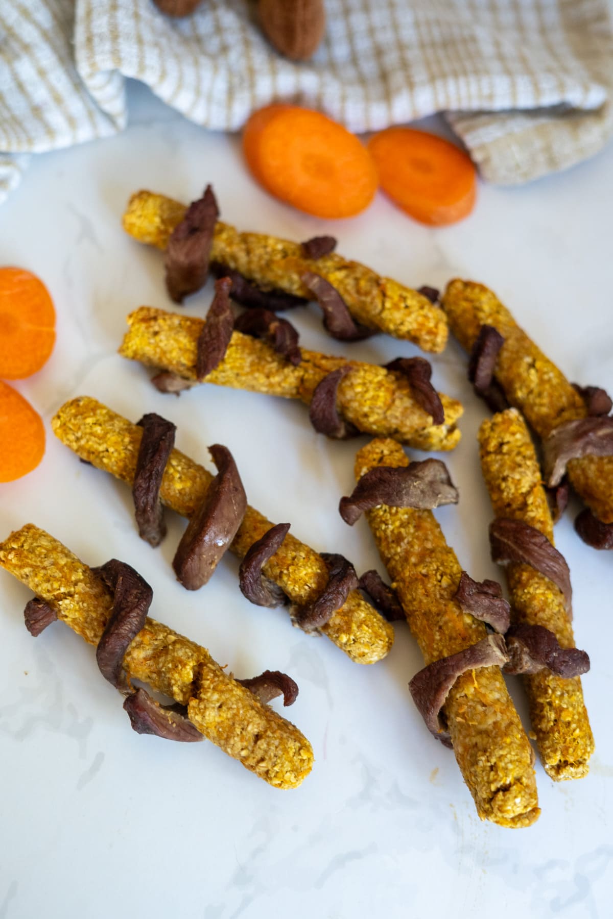 Several homemade duck dog treats made of orange sticks wrapped with strips of dried meat are arranged on a white surface, with slices of carrots and a cloth in the background.